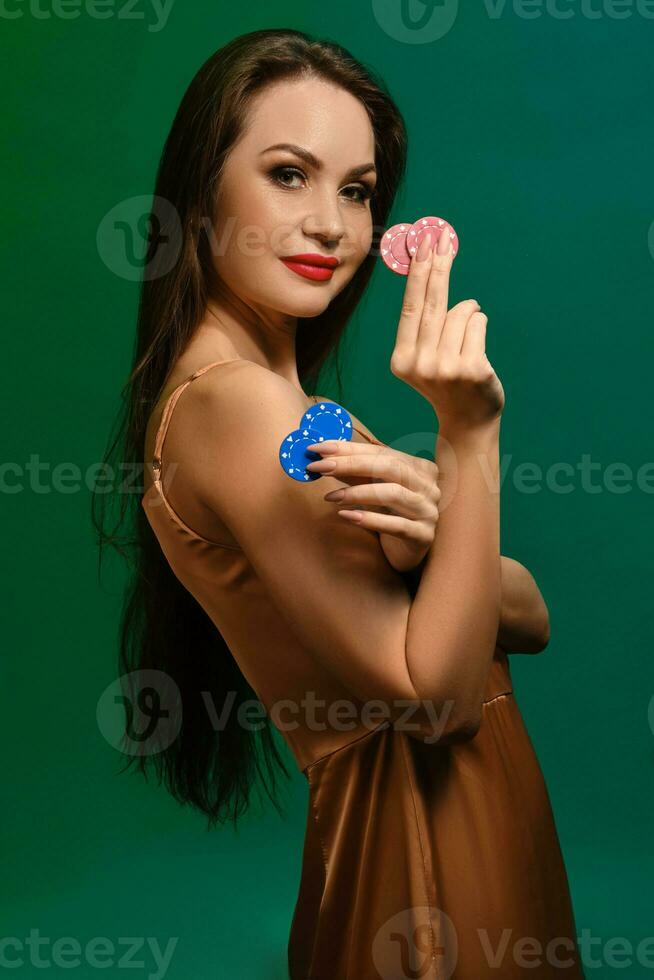 brunetta ragazza nel seta beige vestire. sorridente, mostrando quattro colorato patatine fritte, in posa lateralmente su verde sfondo. poker, casinò. avvicinamento foto