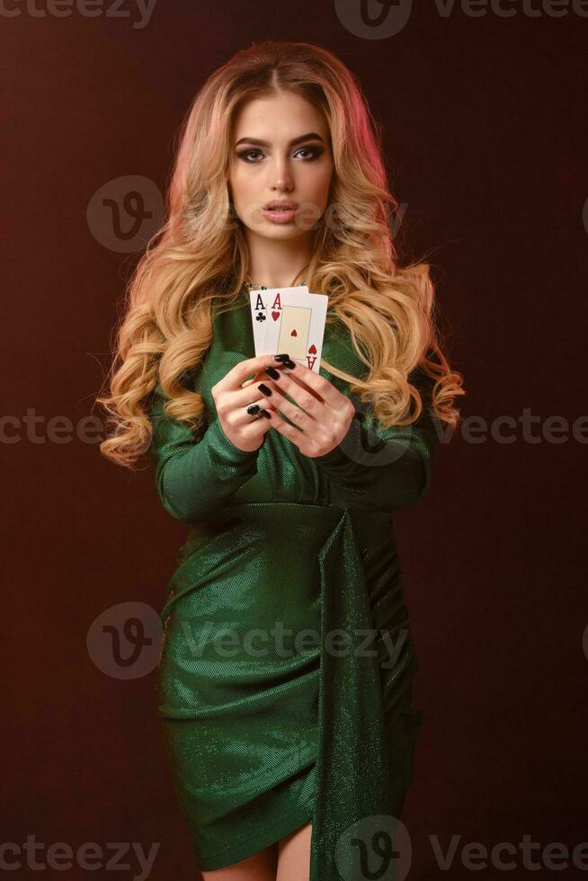 bionda Riccio ragazza nel verde elegante vestito e gioielleria. lei è mostrando Due giocando carte, in posa su Marrone sfondo. poker, casinò. avvicinamento foto