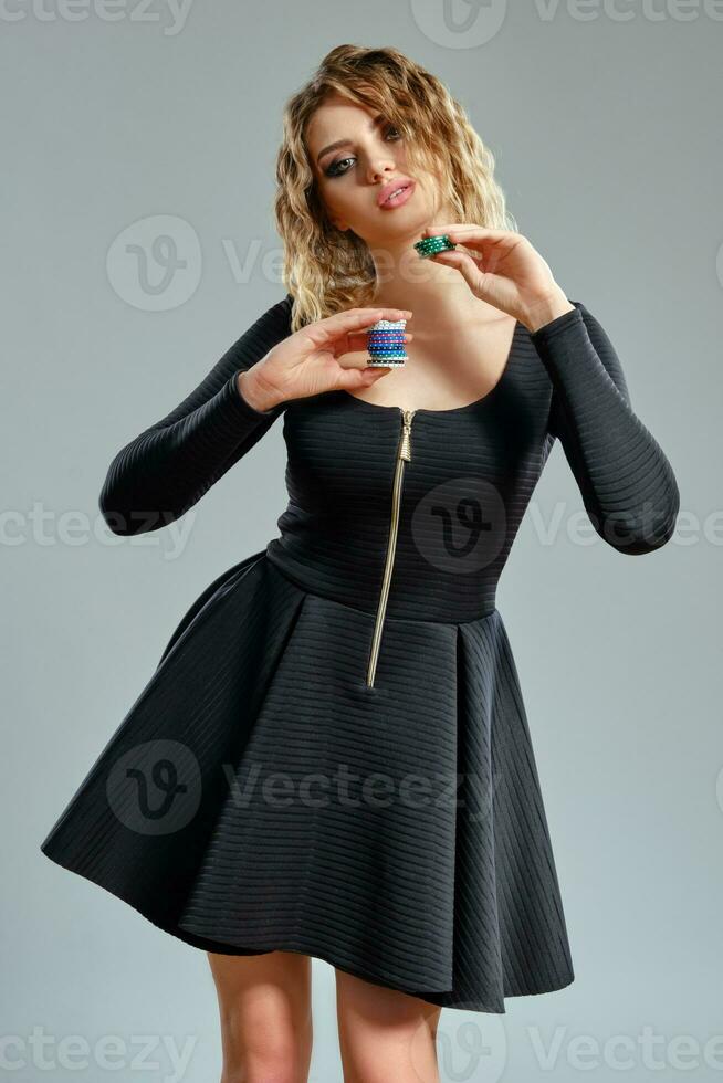 bionda femmina nel nero corto vestito mostrando alcuni colorato patatine fritte, in posa contro grigio sfondo. gioco d'azzardo intrattenimento, poker, casinò. avvicinamento. foto