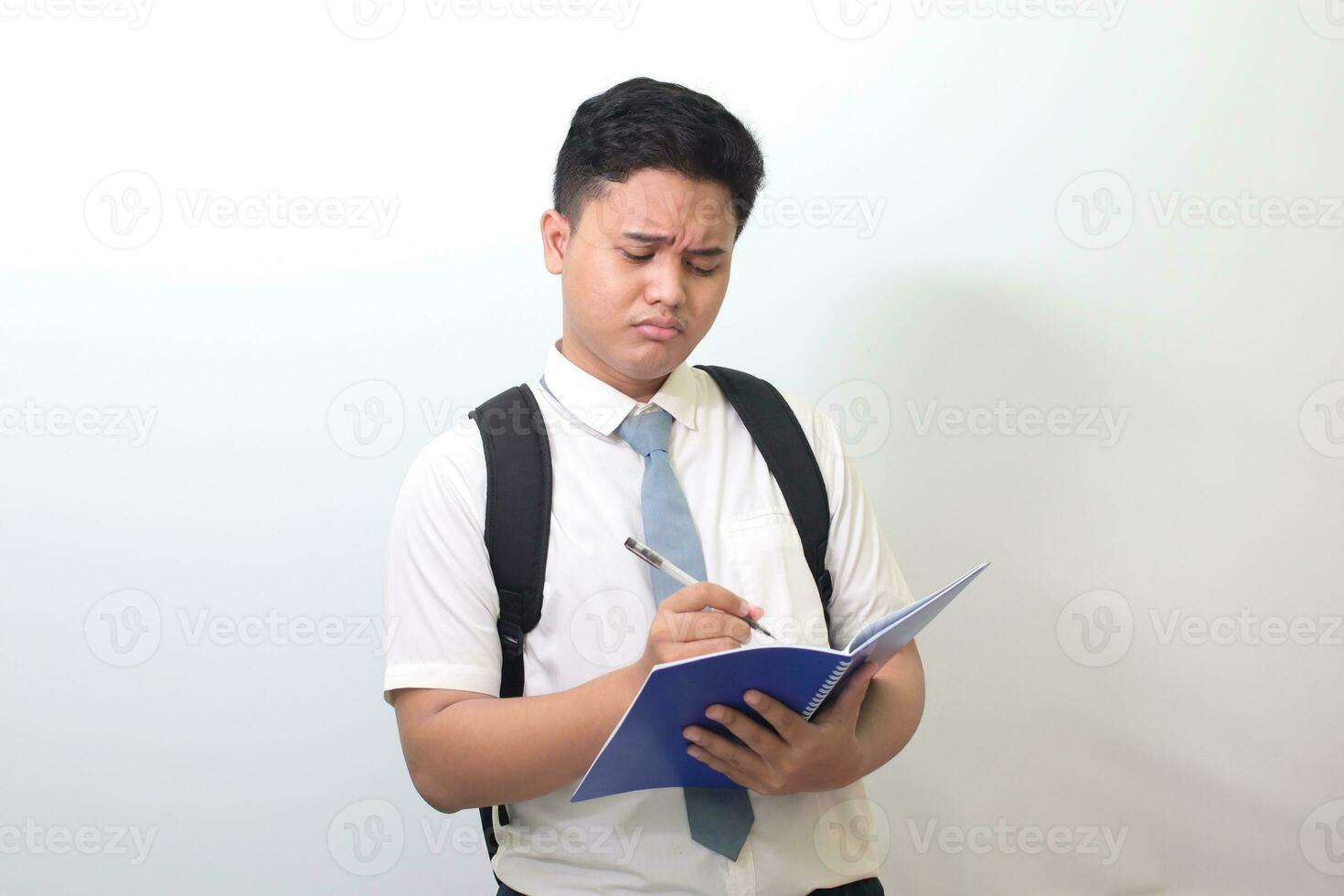 indonesiano anziano alto scuola alunno indossare bianca camicia uniforme con grigio cravatta scrittura su Nota libro utilizzando penna con infastidito e frustrato espressione. isolato Immagine su bianca sfondo foto
