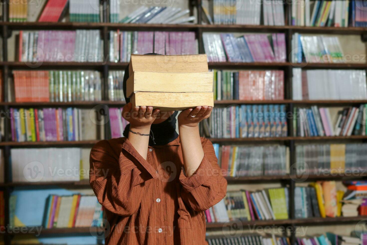 ritratto di asiatico hijab donna copertura sua testa con un' mucchio di libri nel davanti di biblioteca scaffale. musulmano ragazza lettura un' libro. concetto di alfabetizzazione e conoscenza foto