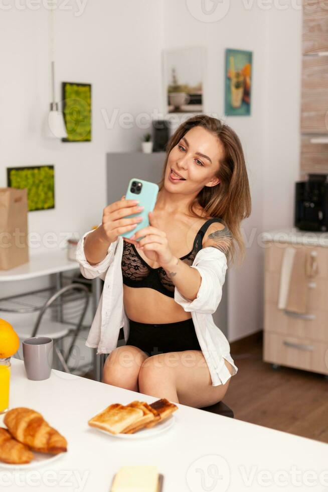 donna godendo assunzione autoscatto nel casa cucina utilizzando smartphone. seducente donna con tatuaggi utilizzando Telefono indossare tempra biancheria intima nel il mattina. foto