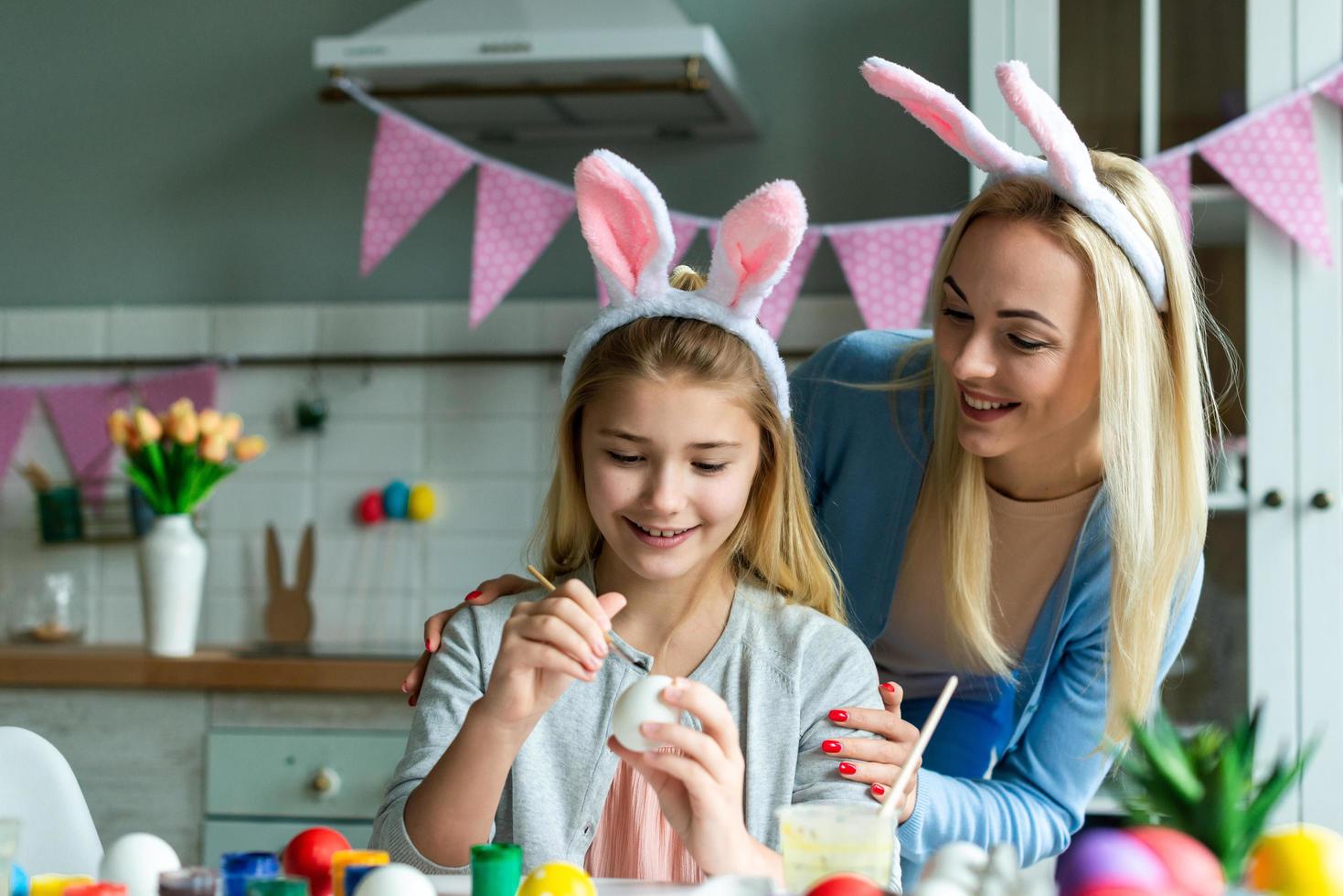 divertente, allegro, gioiosa mamma che insegna, allenandola carina, carina, piccola, piccola figlia disegna, dipingere, decorare le uova di pasqua, insieme indossando orecchie da coniglio, preparando per la pasqua foto