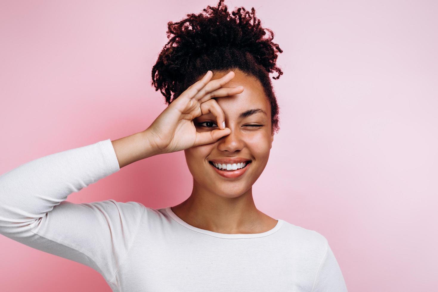 ragazza con una pelle morbida, liscia e pulita, sorriso raggiante, gesticolando segno ok vicino a un occhio isolato su sfondo rosa foto