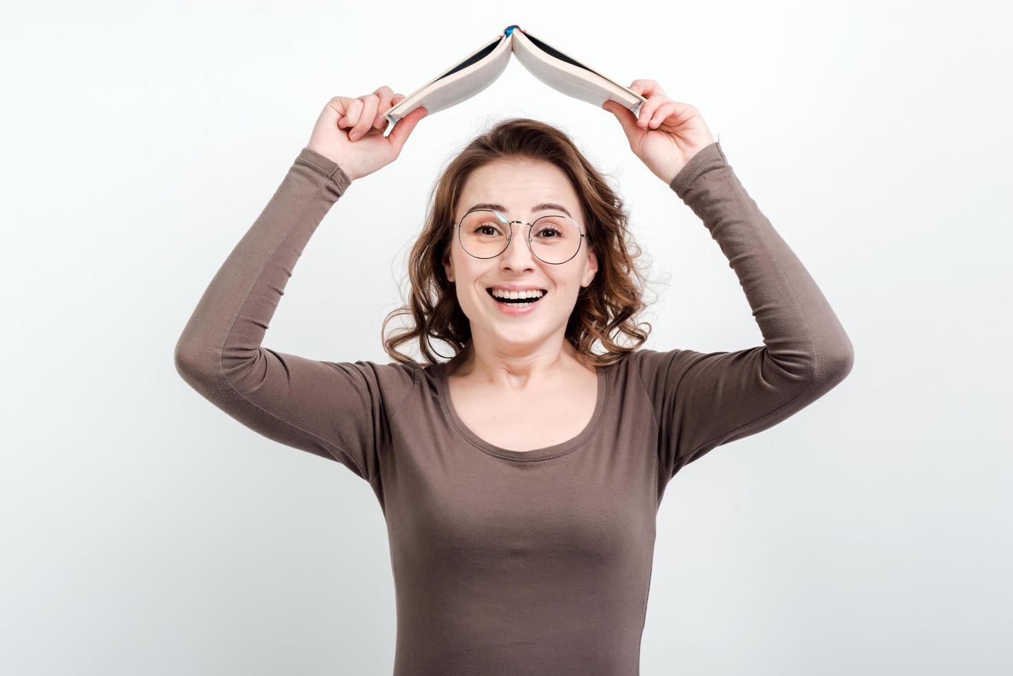 felice e divertente donna con gli occhiali in piedi su uno sfondo di studio con in mano un libro sopra la sua testa foto