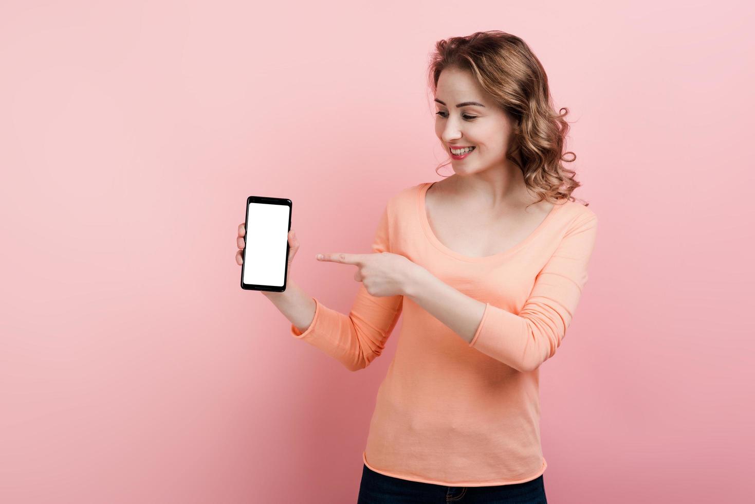 una ragazza sorridente tiene in mano un telefono e punta il dito su uno schermo bianco. isolato su uno sfondo rosa. foto