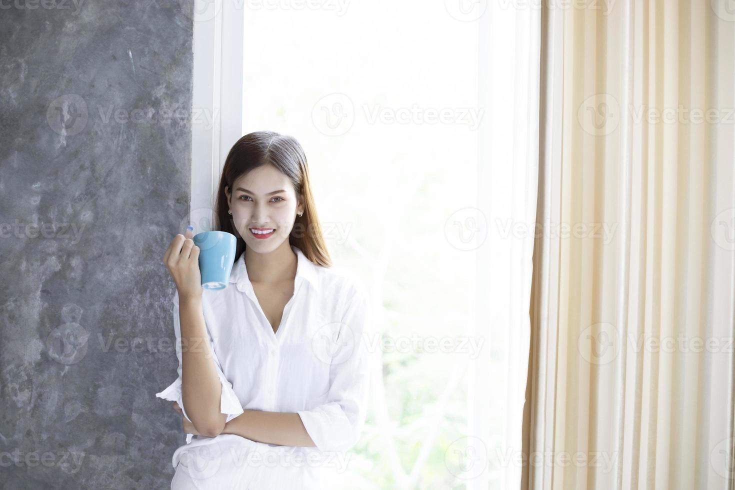 donne asiatiche che bevono caffè e si svegliano nel suo letto completamente riposate e aprono le tende al mattino per prendere aria fresca al sole foto
