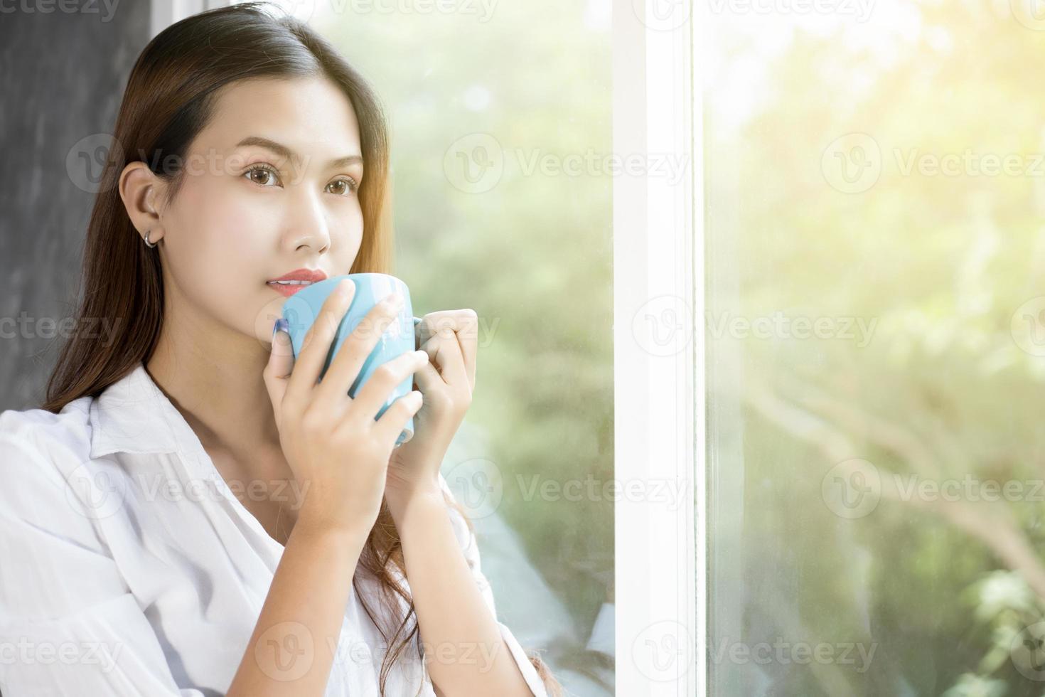 donne asiatiche che bevono caffè e si svegliano nel suo letto completamente riposate e aprono le tende al mattino per prendere aria fresca al sole foto