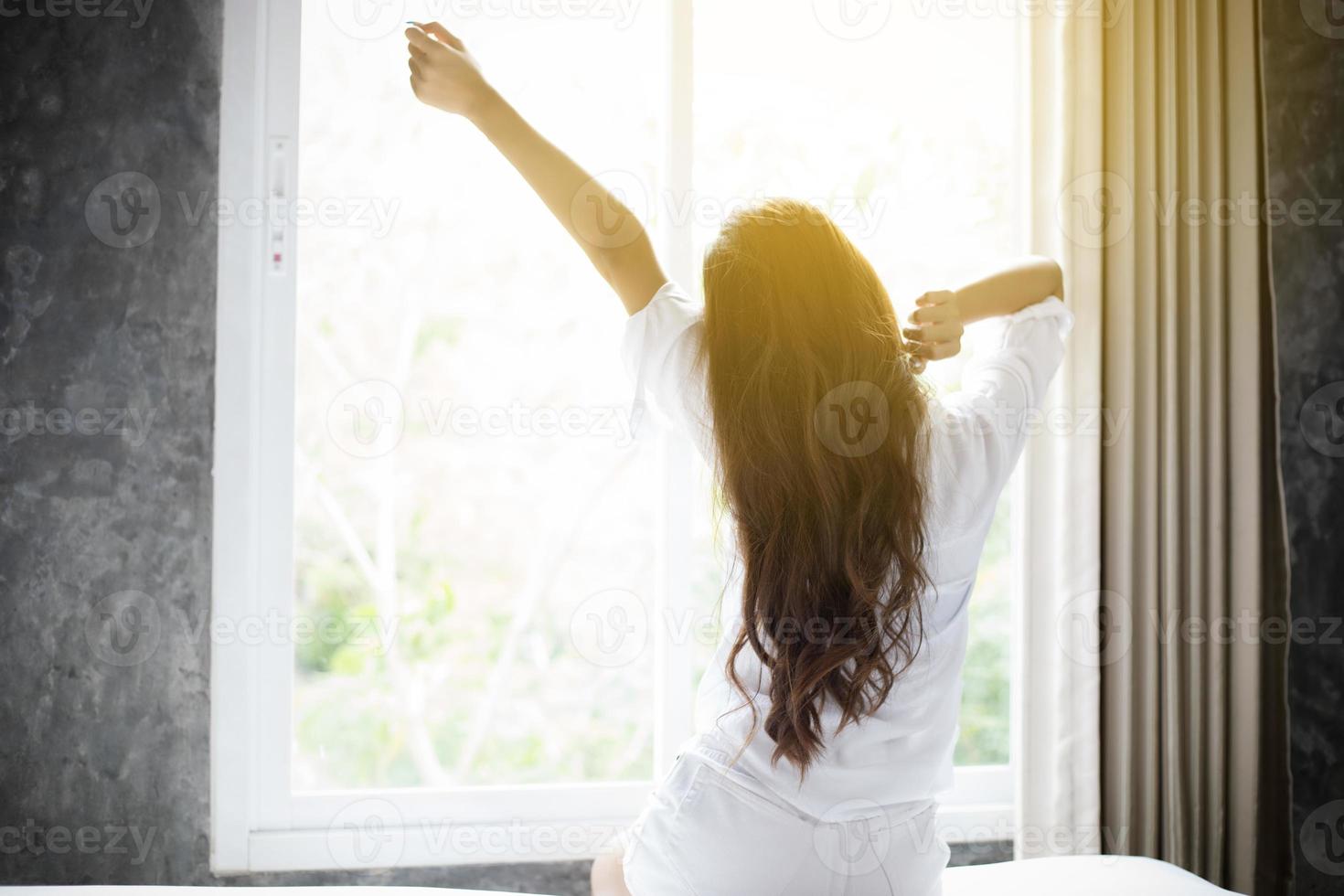 donna asiatica bella giovane donna sorridente seduta sul letto e stretching la mattina in camera da letto dopo essersi svegliata nel suo letto completamente riposata e aprire le tende al mattino per prendere aria fresca. foto