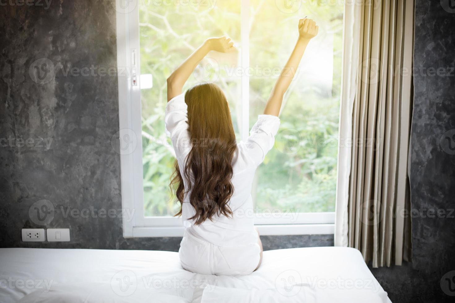 donna asiatica bella giovane donna sorridente seduta sul letto e stretching la mattina in camera da letto dopo essersi svegliata nel suo letto completamente riposata e aprire le tende al mattino per prendere aria fresca. foto