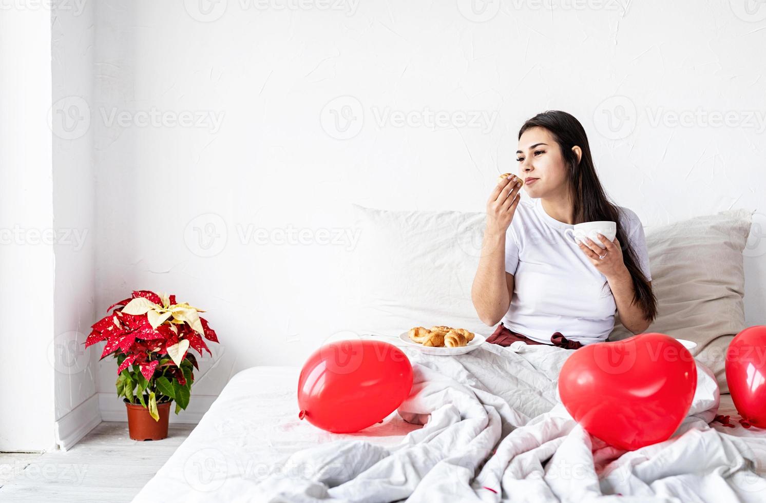 giovane donna bruna seduta sveglia nel letto con palloncini e decorazioni a forma di cuore rosso che bevono caffè mangiando croissant foto