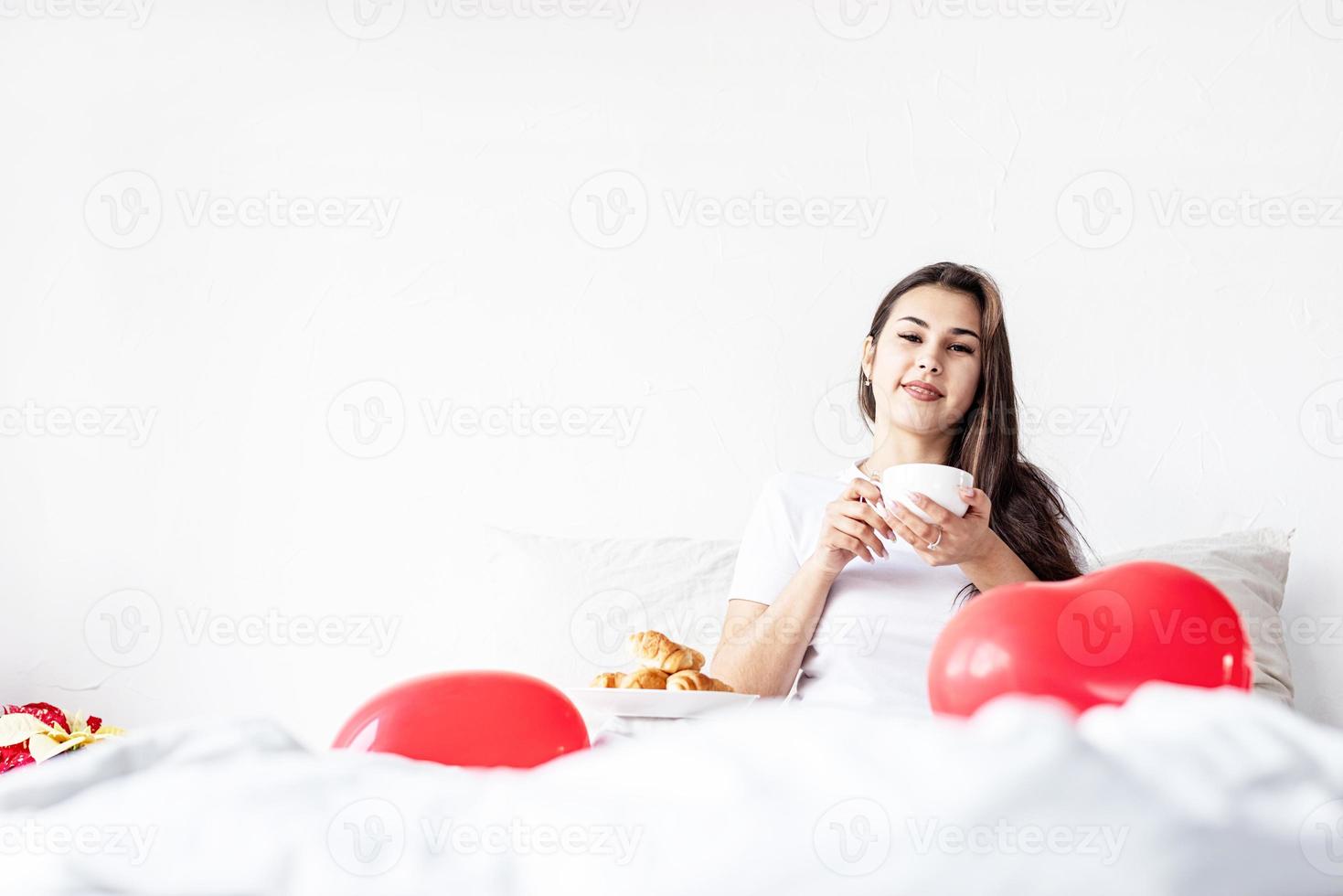 giovane donna bruna seduta sveglia nel letto con palloncini e decorazioni a forma di cuore rosso che bevono caffè mangiando croissant foto