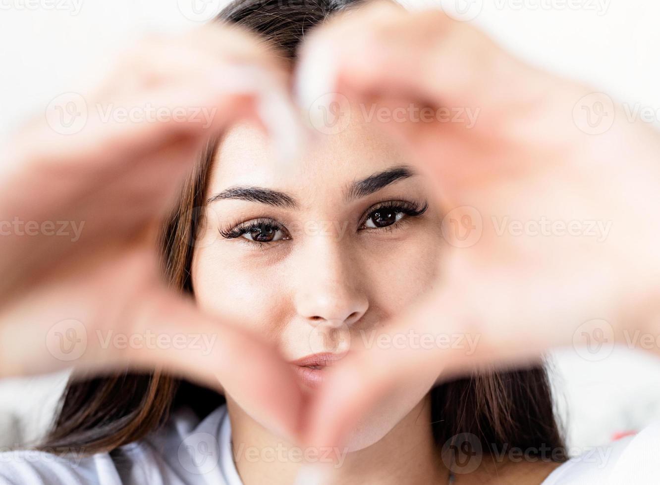 giovane donna bruna felice in maglietta bianca che mostra il segno del cuore con le mani davanti al viso foto