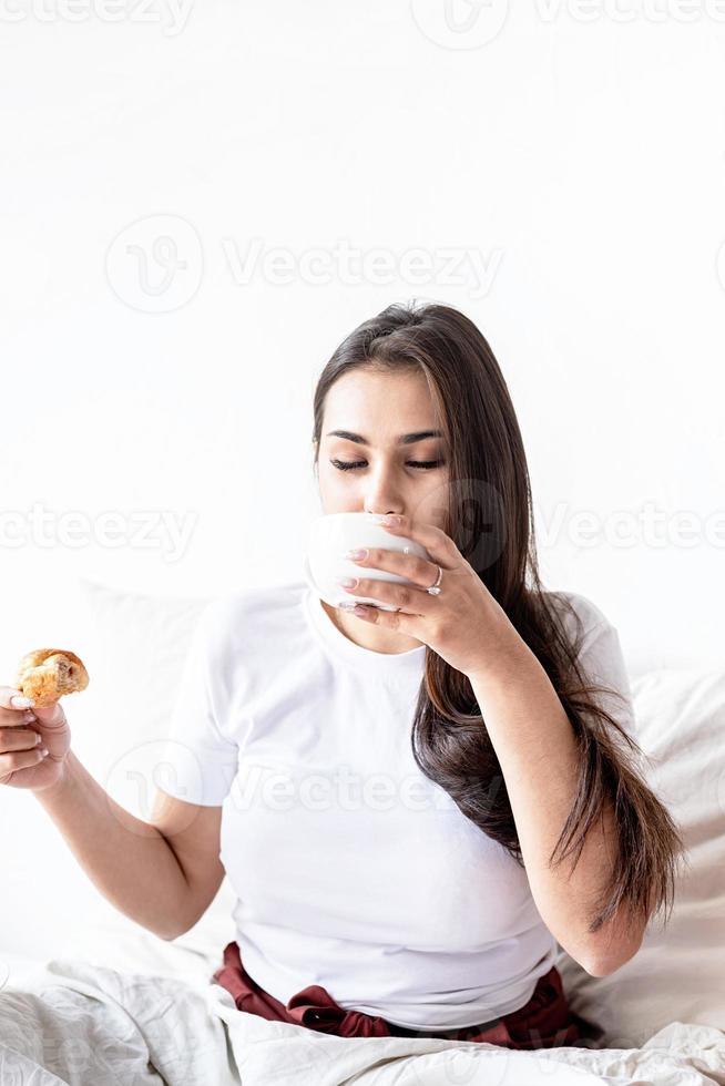 giovane donna bruna seduta nel letto con mangiare croissant foto
