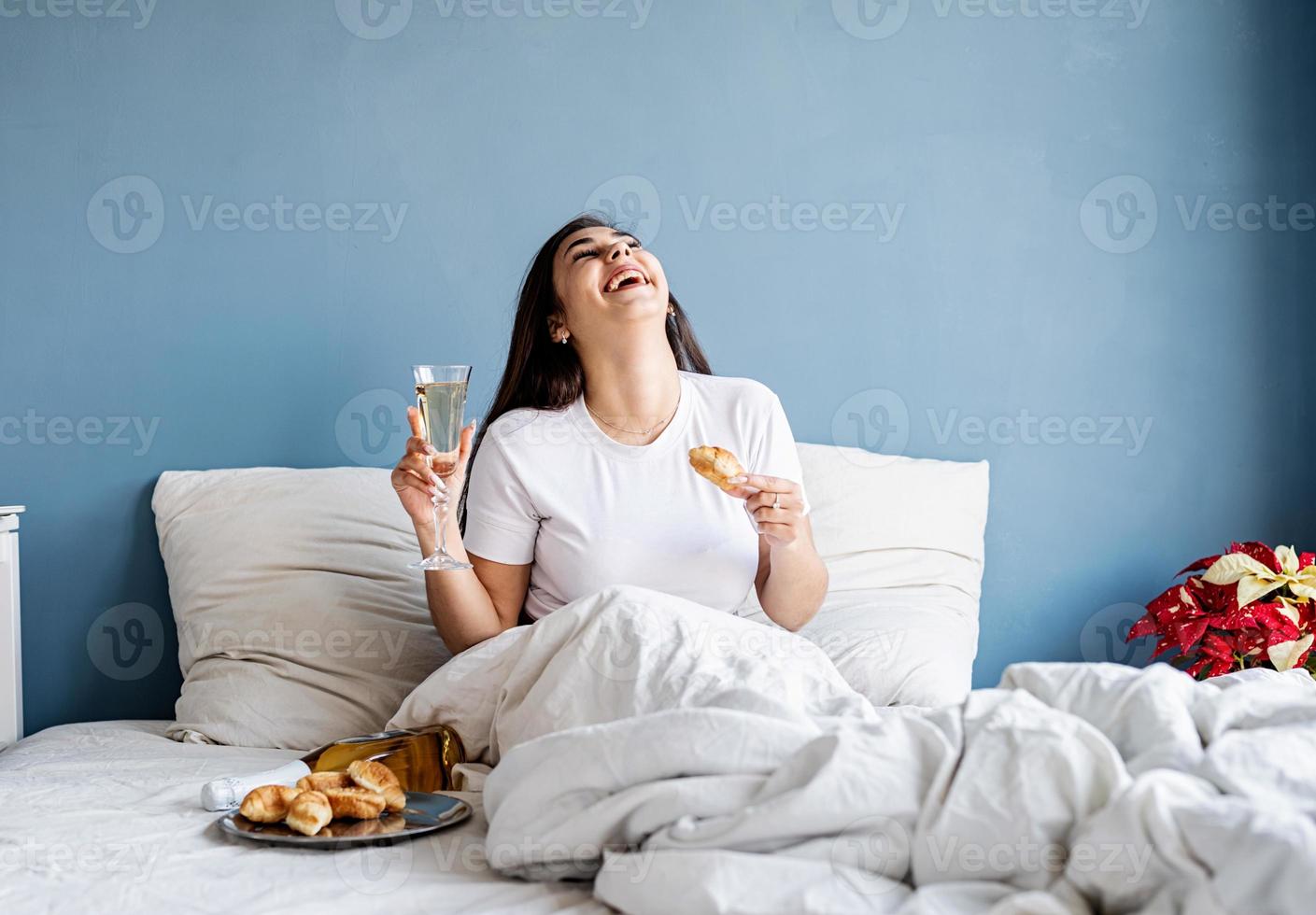 giovane donna bruna seduta sveglia nel letto con palloncini e decorazioni a forma di cuore rosso bevendo champagne mangiando croissant foto