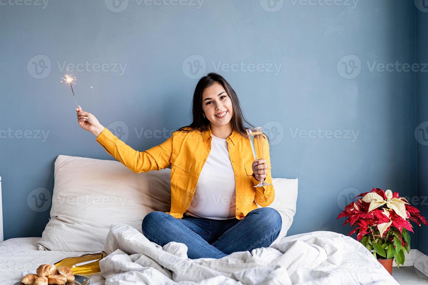 giovane donna bruna seduta nel letto a bere champagne con in mano un sparkler foto