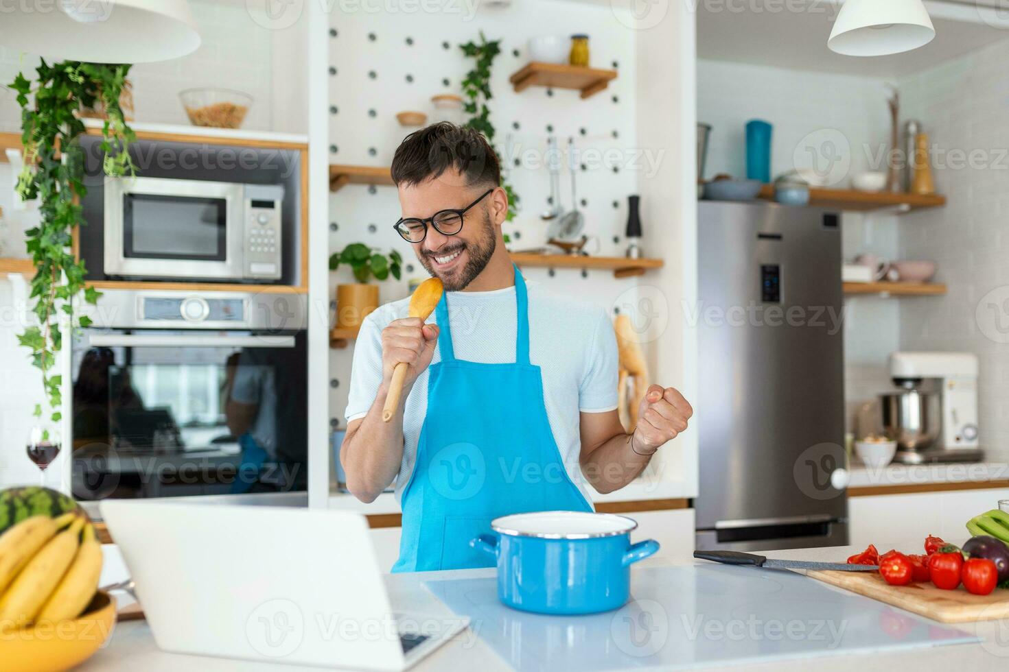 divertente giovane uomo cantando canzone in frusta, cucinando nel moderno cucina, contento maschio Tenere utensili da cucina come microfono, ascoltando per musica, ballare, fare lavori di casa a casa, preparazione prima colazione foto