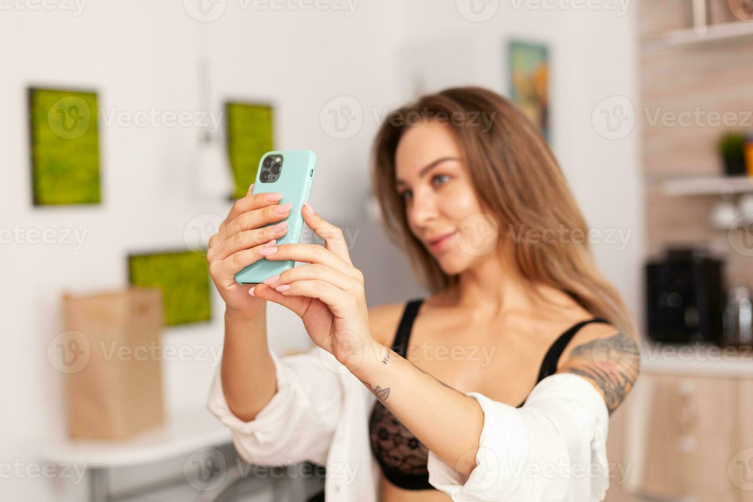 ritratto di bellissimo donna guardare a Telefono telecamera mentre assunzione autoscatto nel casa cucina wering sexy biancheria intima. seducente donna con tatuaggi utilizzando smartphone indossare tempra biancheria intima nel il mattina. foto