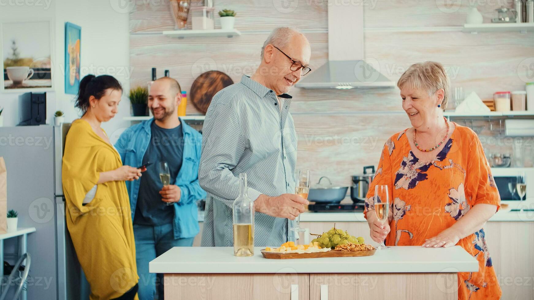 allegro vecchio anziano adulto famiglia abbracciare ridendo danza potabile un' bicchiere di bianca vino nel davanti giovane coppia. nel il cucina. nonna e Nonno Abbracciare una persona sorridente e figlia assunzione un' foto
