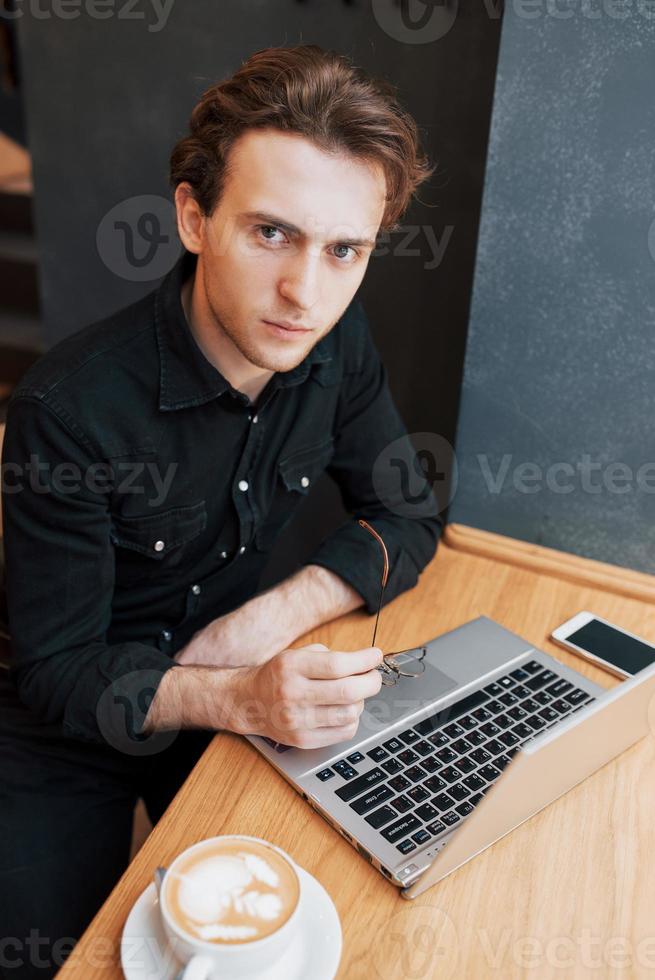 designer uomo creativo che lavora sul suo computer portatile mentre aspetta gli ordini al suo bar preferito al chiuso, studente maschio che lavora su net-book durante la colazione mattutina nell'interno moderno della caffetteria foto