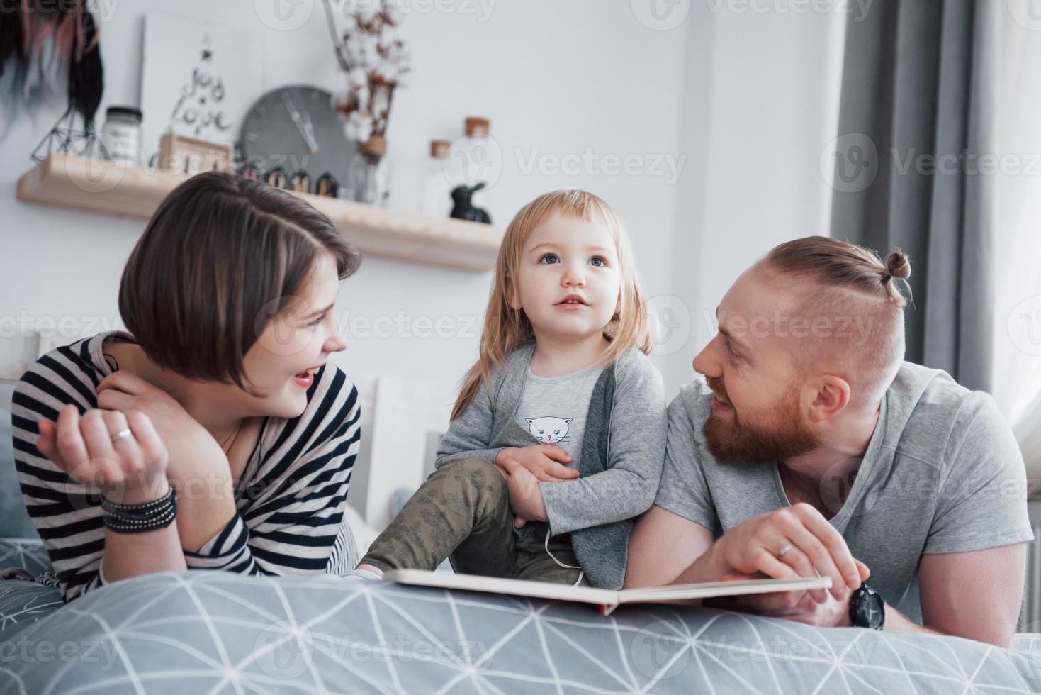 padre, madre e figlia piccola che leggono un libro per bambini su un divano in soggiorno. la grande famiglia felice ha letto un libro interessante in un giorno festivo. i genitori amano i loro figli foto