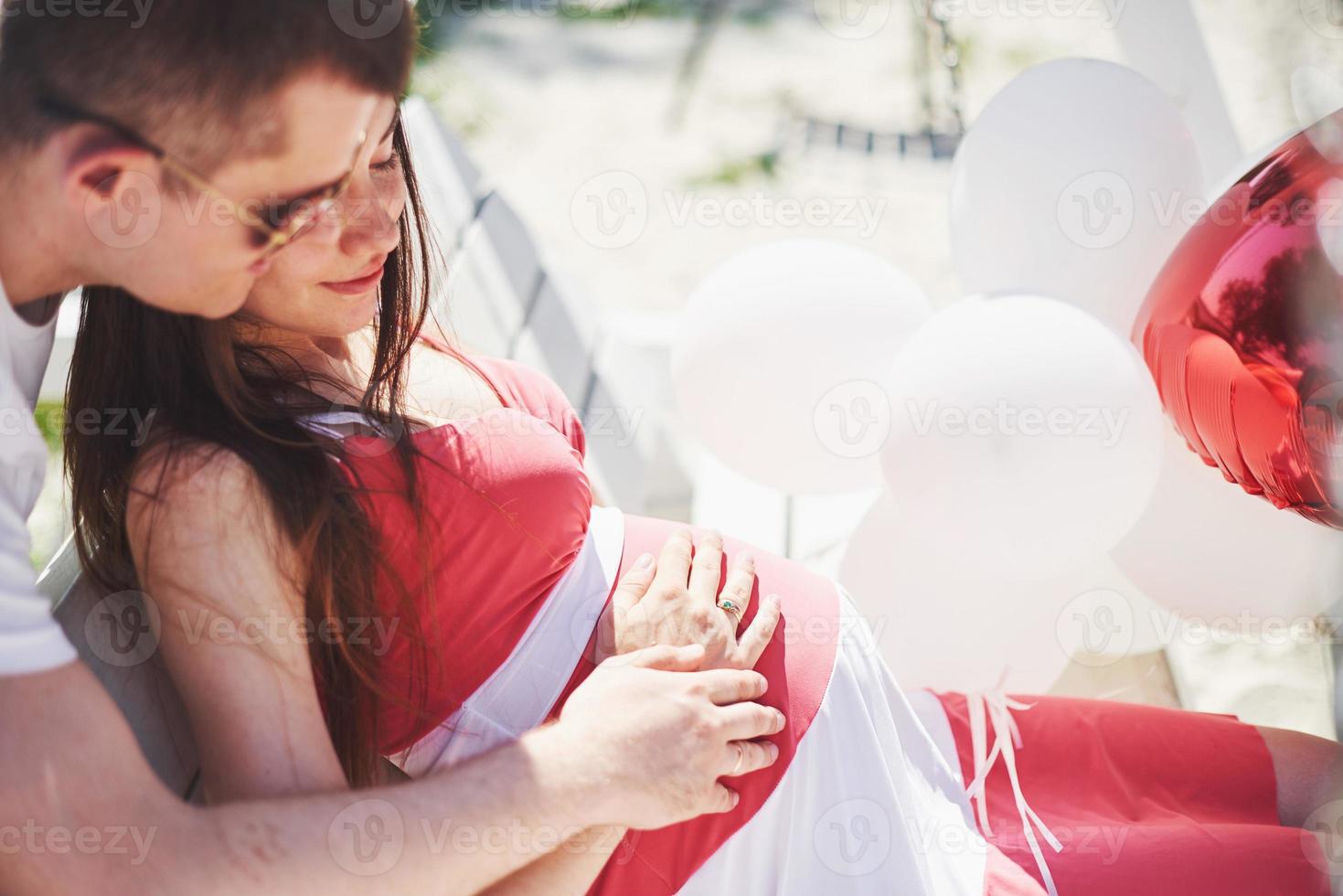 aspettando bambino. donna incinta con l'amato marito si siede su una panchina. la mano del marito abbraccia una moglie a pancia rotonda. paternità. donna incinta in un abito bianco in ciliegio con un fiocco rosso. avvicinamento. Nove mesi foto