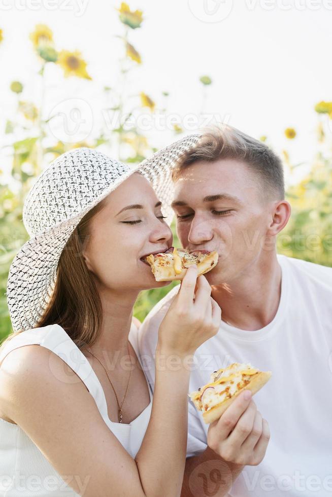 giovane coppia che fa picnic sul campo di girasoli al tramonto, mordendo un pezzo di pizza insieme foto