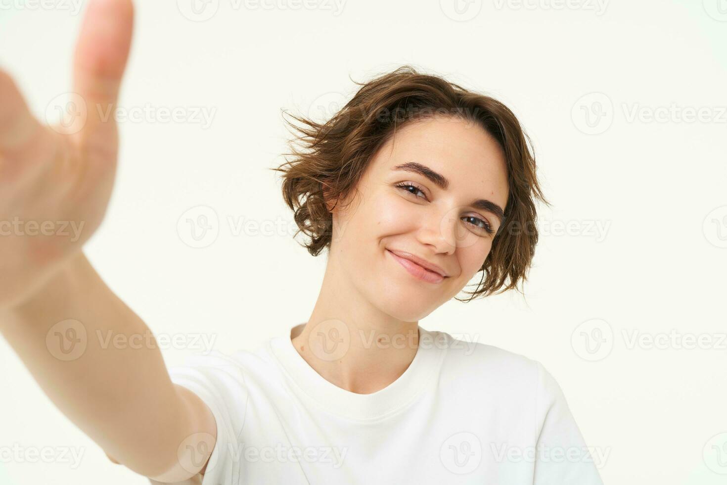 stile di vita e le persone. giovane donna sorridente e assunzione autoscatto, in posa per foto, Tenere telecamera con uno mano, in piedi al di sopra di bianca sfondo foto