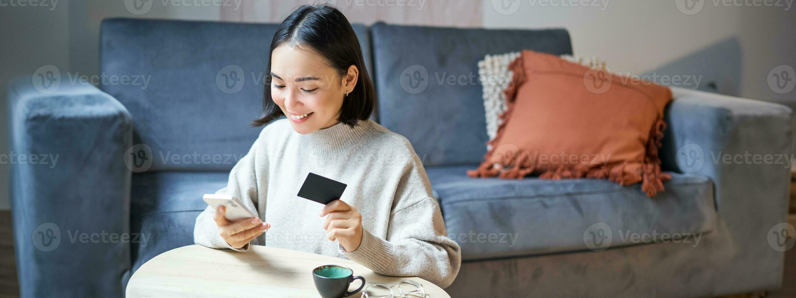 sorridente carino asiatico donna utilizzando credito carta e smartphone, pagare fatture in linea, Tenere mobile Telefono, guardare a telecamera foto