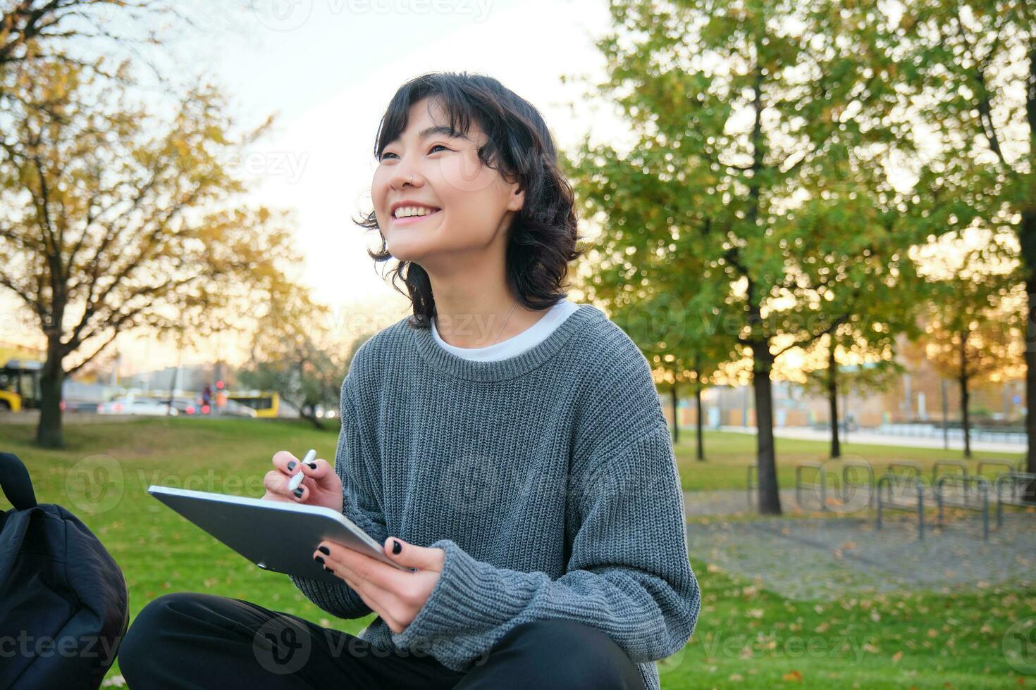 giovane coreano donna si siede nel parco, disegna su digitale tavoletta, scrive nel aggeggio con penna attrezzo, sembra in giro e fa Appunti, crea opera d'arte o graffi foto