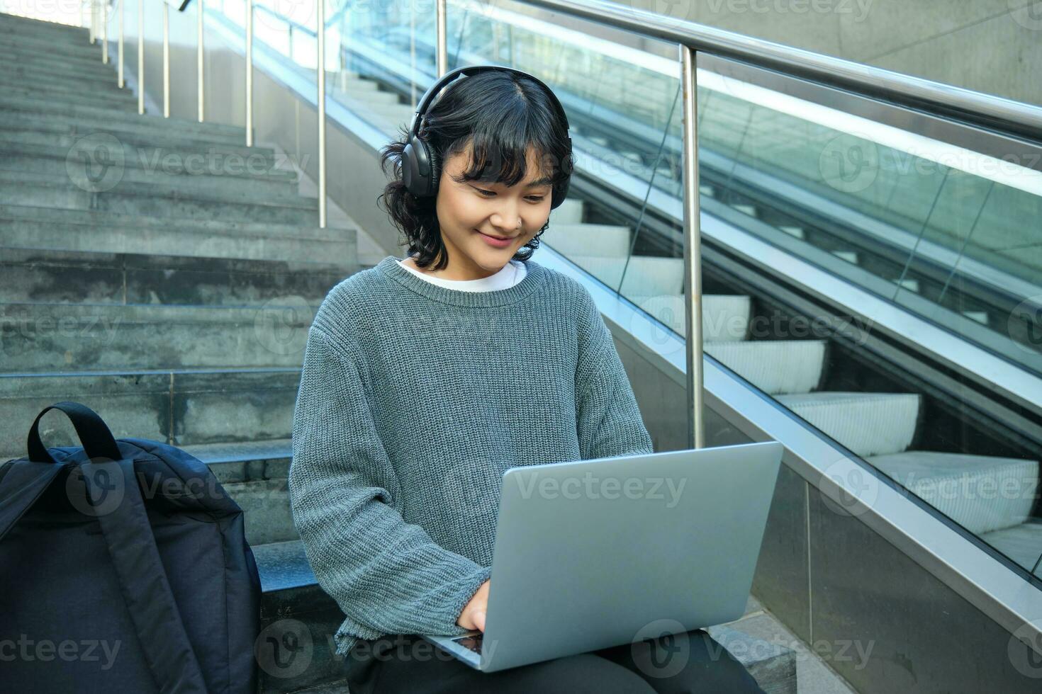 ritratto di bellissimo giovane coreano donna, alunno si siede su le scale nel pubblico posto, ascolta musica nel cuffia, lavori su progetto su computer portatile, lavori remoto foto