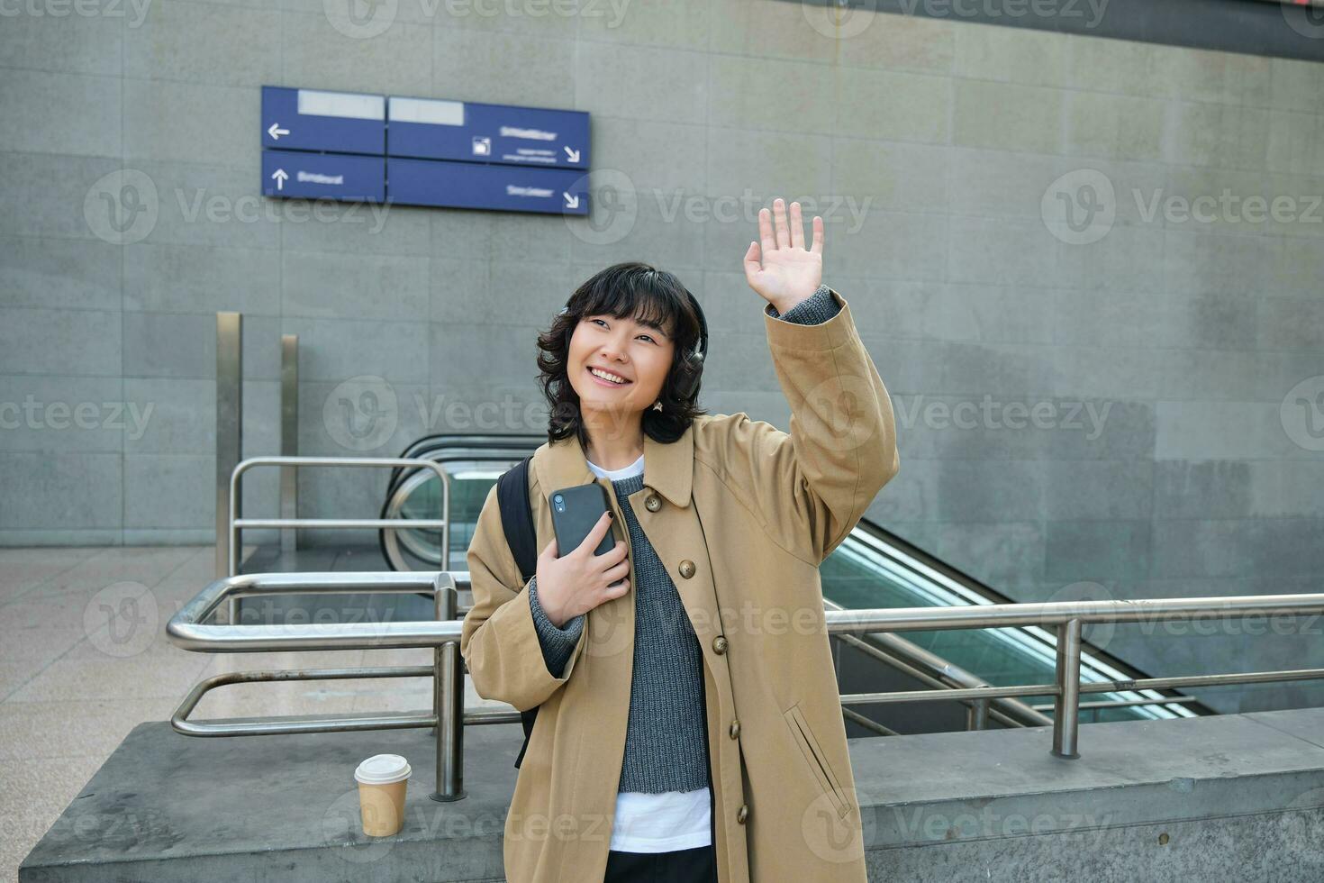ritratto di carino sorridente asiatico ragazza nel cuffia, sta su strada con smartphone, onde mano a qualcuno, dice ciao, saluta amico foto