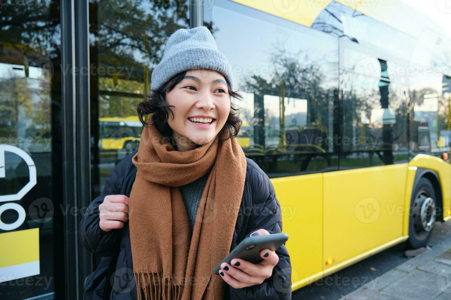 ritratto di ragazza in piedi vicino autobus su un' fermare, in attesa per sua pubblico trasporto, controlli programma su smartphone applicazione, detiene mobile Telefono, indossa caldo Abiti foto