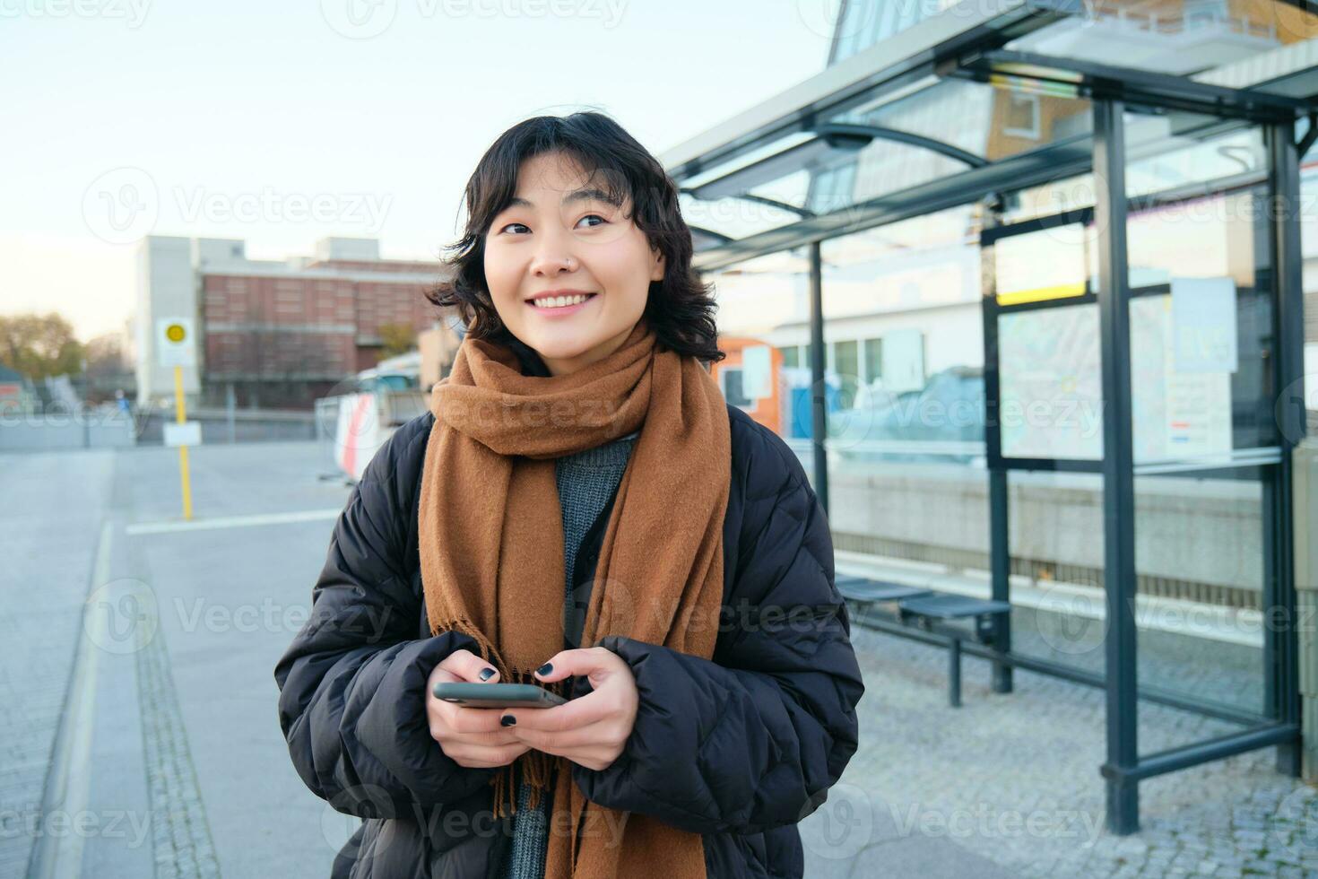 contento sorridente coreano ragazza, utilizzando mobile Telefono, in piedi su autobus fermare con smartphone, guardare a partenza programma su applicazione, in posa nel inverno Abiti foto