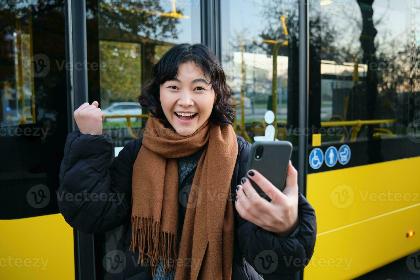 entusiasta asiatico donna, in piedi su autobus fermare con smartphone, guardare a Telefono schermo con stupito, trionfante viso, vincente, sentire grande notizia su video Chiacchierare foto