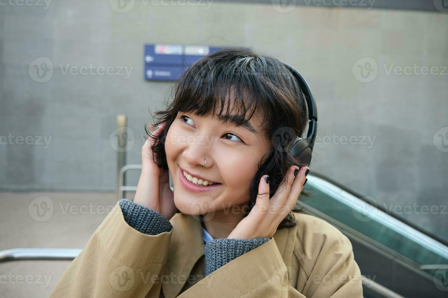 bellissimo sorridente coreano ragazza nel cuffia, chiedendosi in giro cittadina, in piedi su strada e sorridente, ascoltando per musica foto