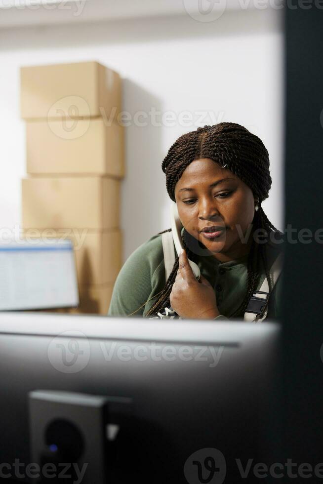 africano americano manager in piedi a contatore scrivania parlando con a distanza cliente utilizzando telefono fisso Telefono, discutere ordine la logistica. magazzino dipendente Lavorando a merce inventario nel magazzino foto