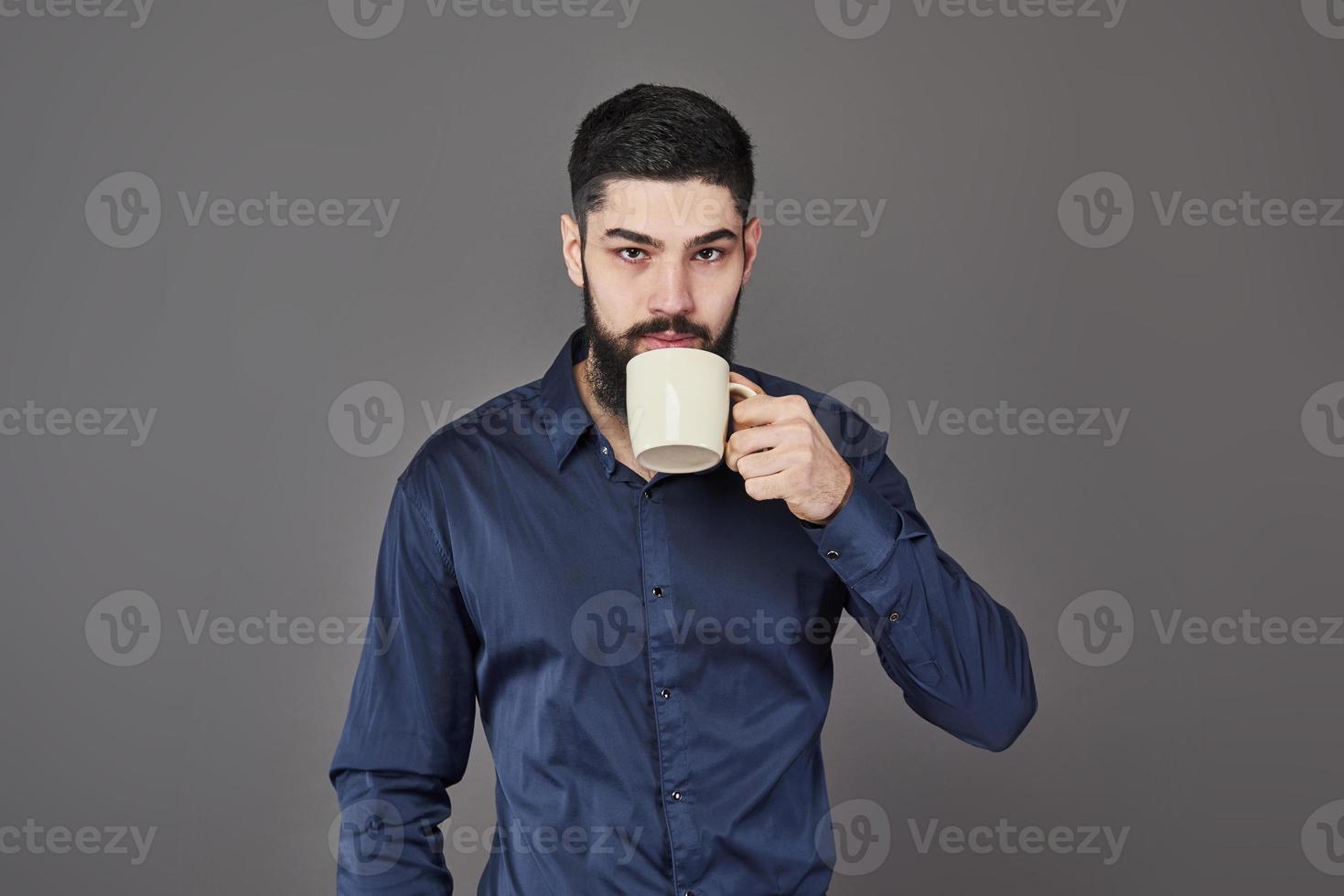 bell'uomo barbuto con capelli alla moda barba e baffi sul viso serio in camicia che tiene tazza bianca o tazza bevendo tè o caffè in studio su sfondo grigio foto