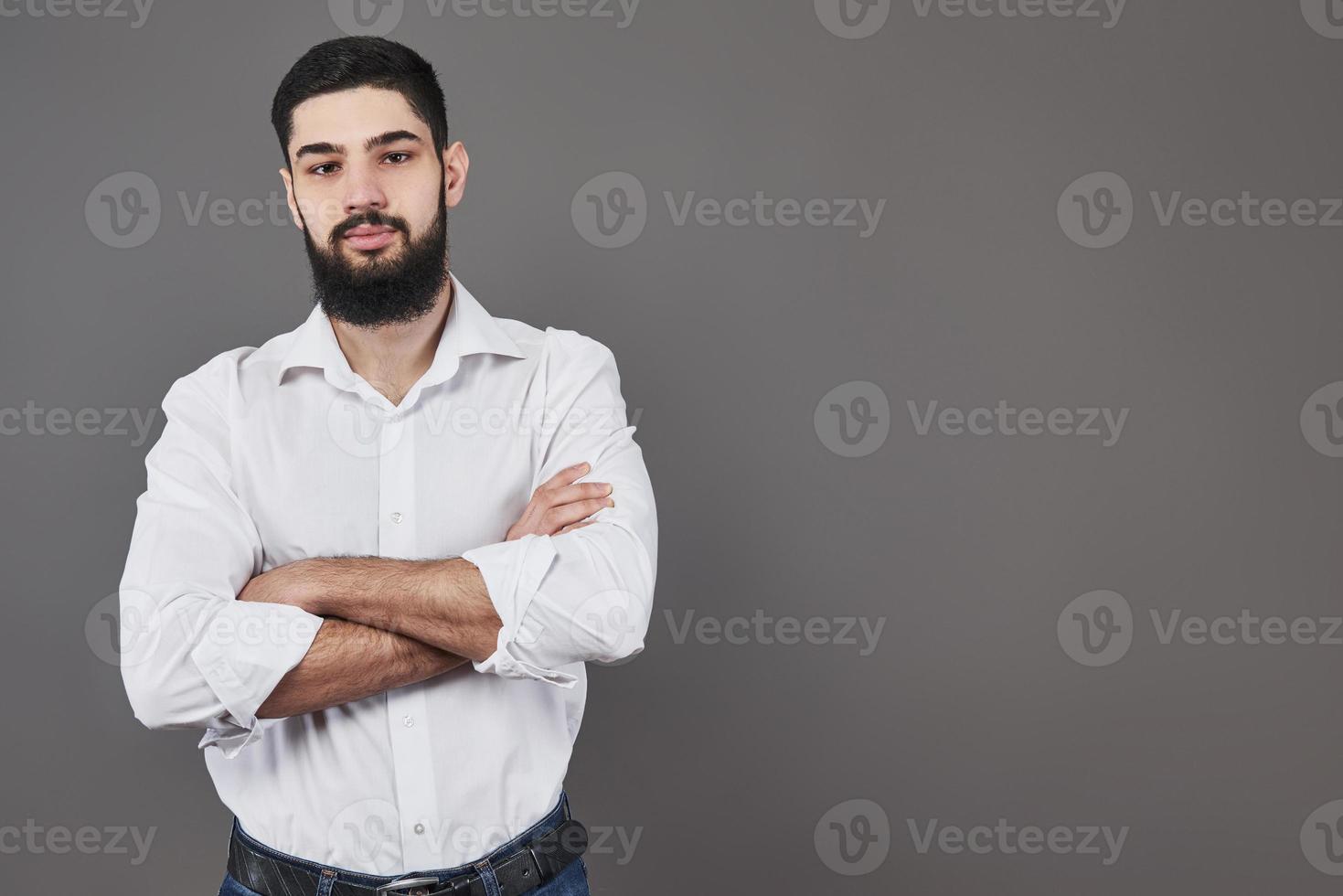 giovane bell'uomo appoggiato al muro grigio con le braccia incrociate. un giovane serio con la barba guarda la telecamera foto