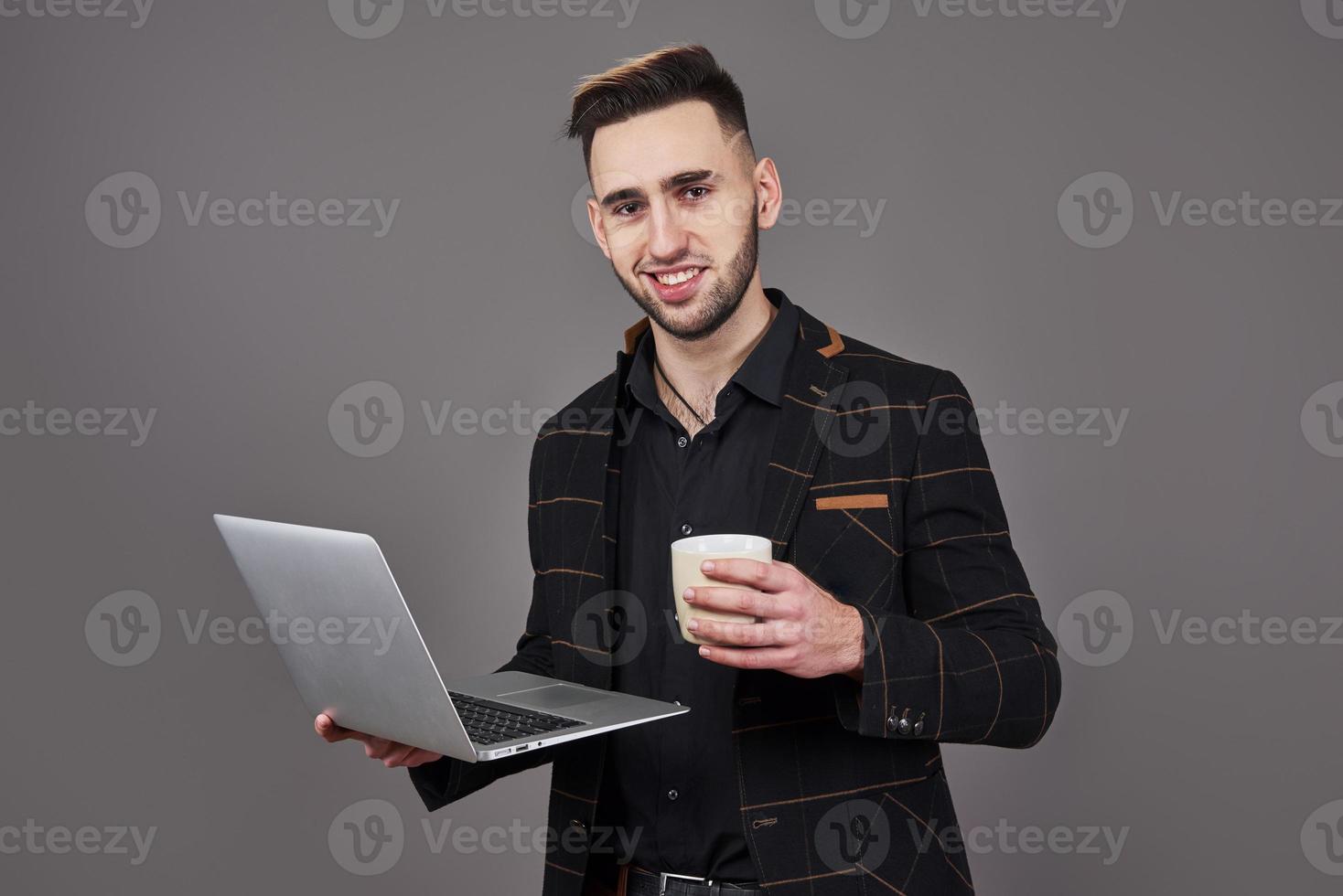 Occupato uomo barbuto in abiti da lavoro che parla tramite smartphone e utilizza il computer portatile mentre si tiene una tazza di caffè in mano su sfondo grigio foto