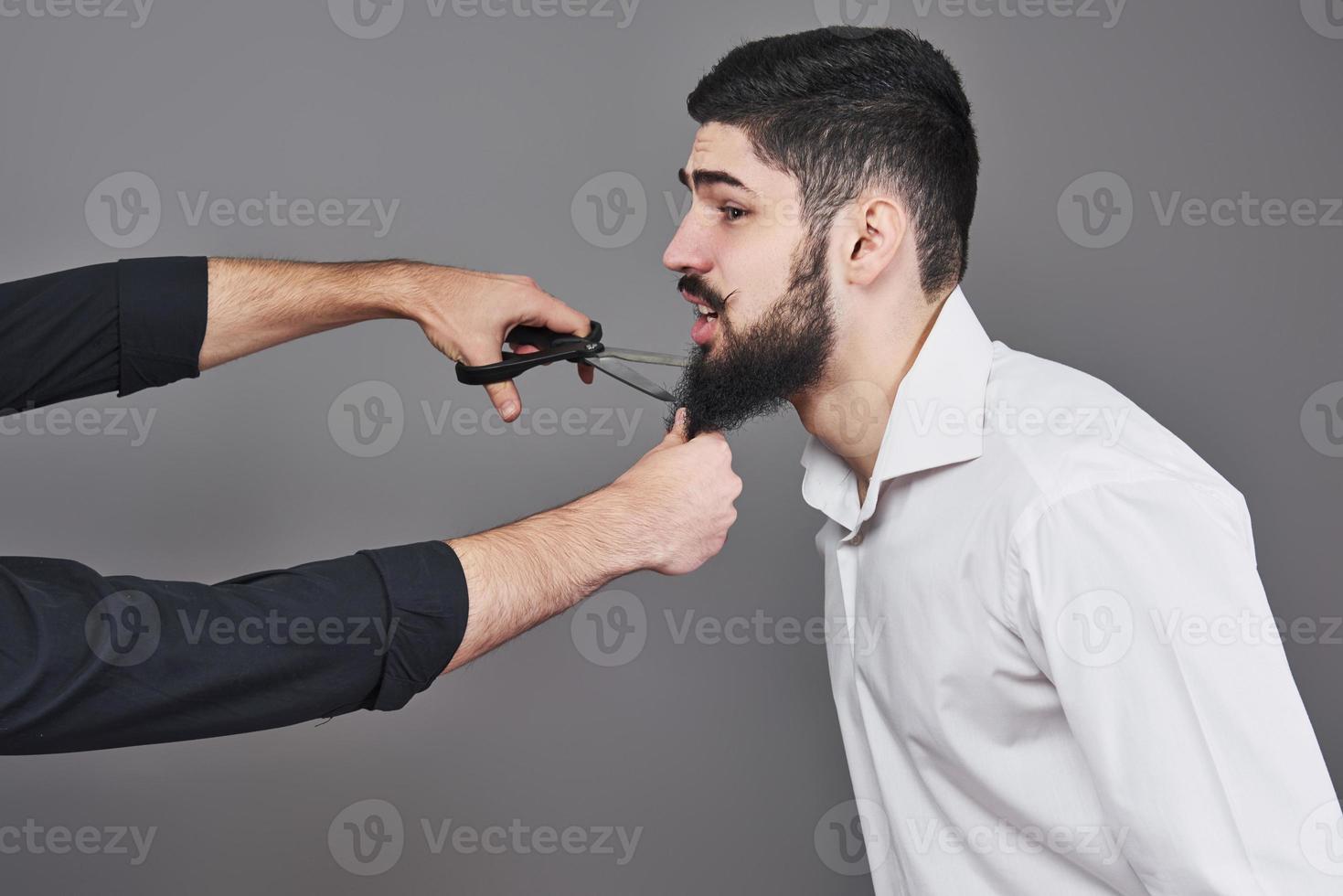 niente più barba. ritratto di un bel giovane che si taglia la barba con le forbici e guarda la telecamera mentre si trova in piedi su uno sfondo grigio. nuova moda foto