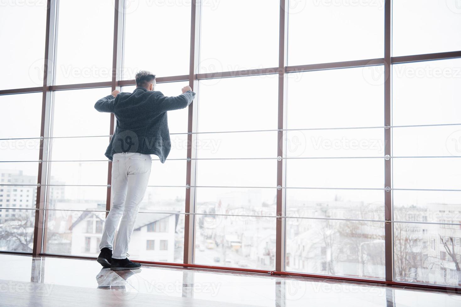 foto a figura intera di un giovane uomo d'affari alla moda che indossa un abito moderno, che è un grande uomo d'azione, in piedi all'ultimo piano di un edificio per uffici che guarda la vista attraverso grandi finestre