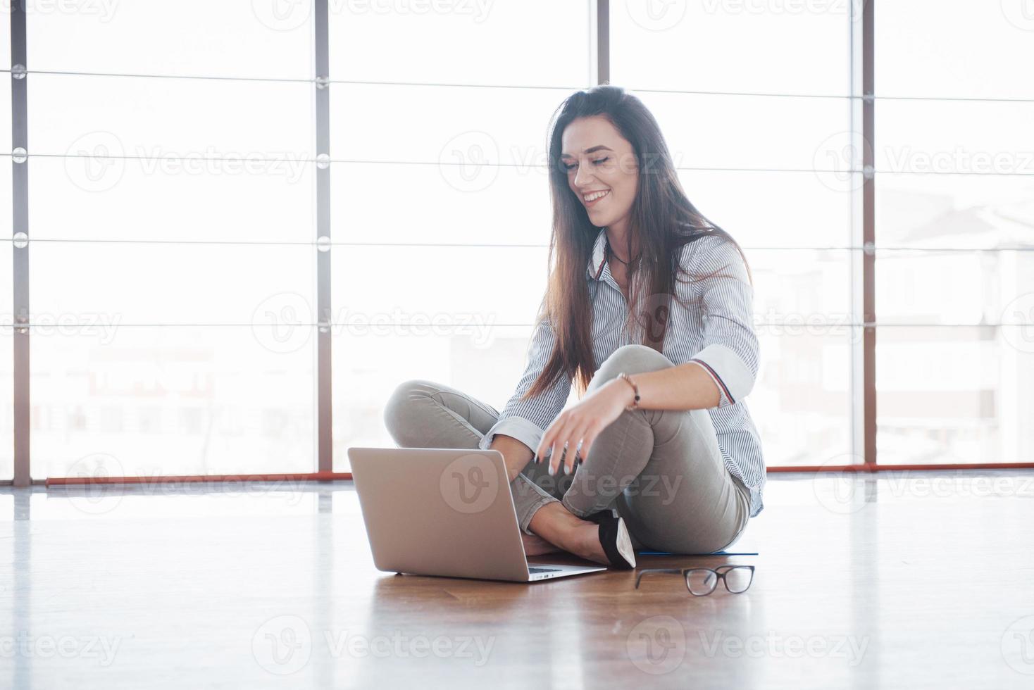 vista laterale di una ragazza attraente che usa un laptop nell'area wifi pubblica e sorride mentre è seduta sul pavimento foto