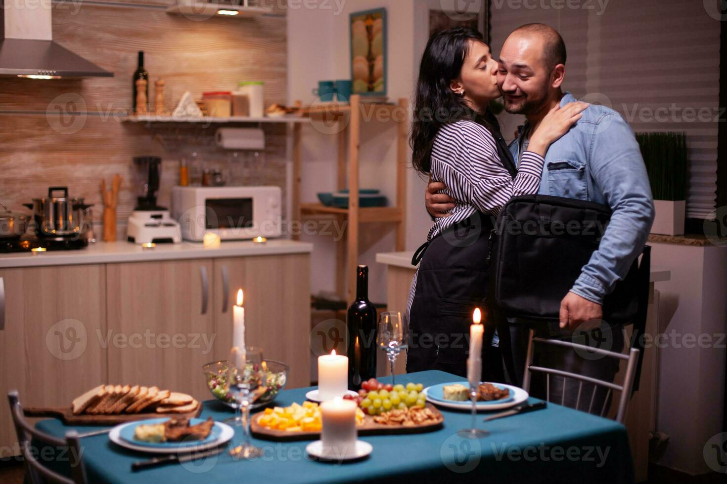 baci marito guancia mentre saluto lui nel cucina per romantico cena. donna preparazione festivo cena con sano cibo, cucinando per il suo uomo romantico cena, parlando, seduta a tavolo foto