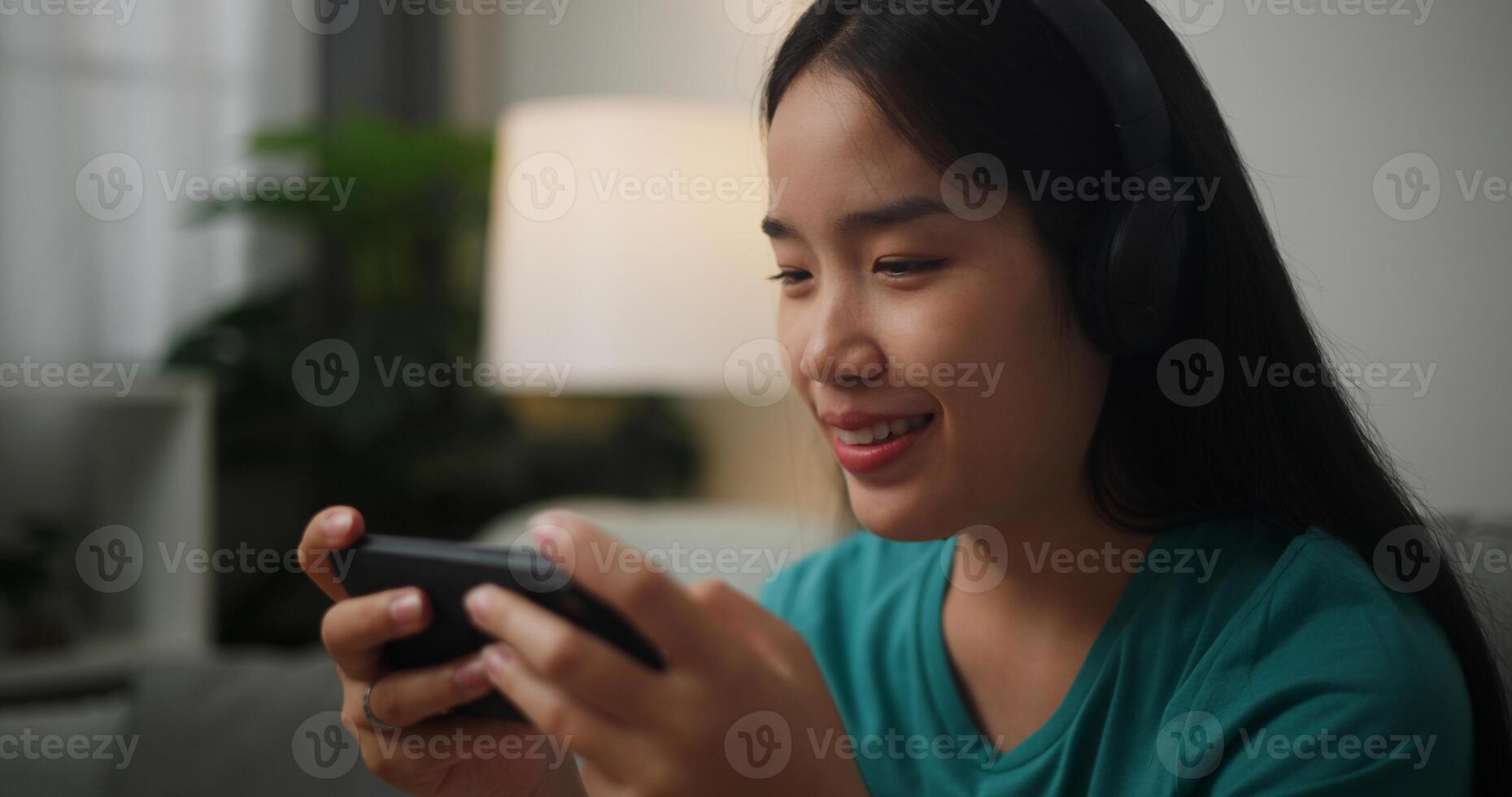 ritratto di giovane asiatico donna indossare bicchieri e cuffie gode giocando in linea esport Giochi su smartphone seduta su divano nel il vivente camera a casa, giocatore stile di vita concetto. foto