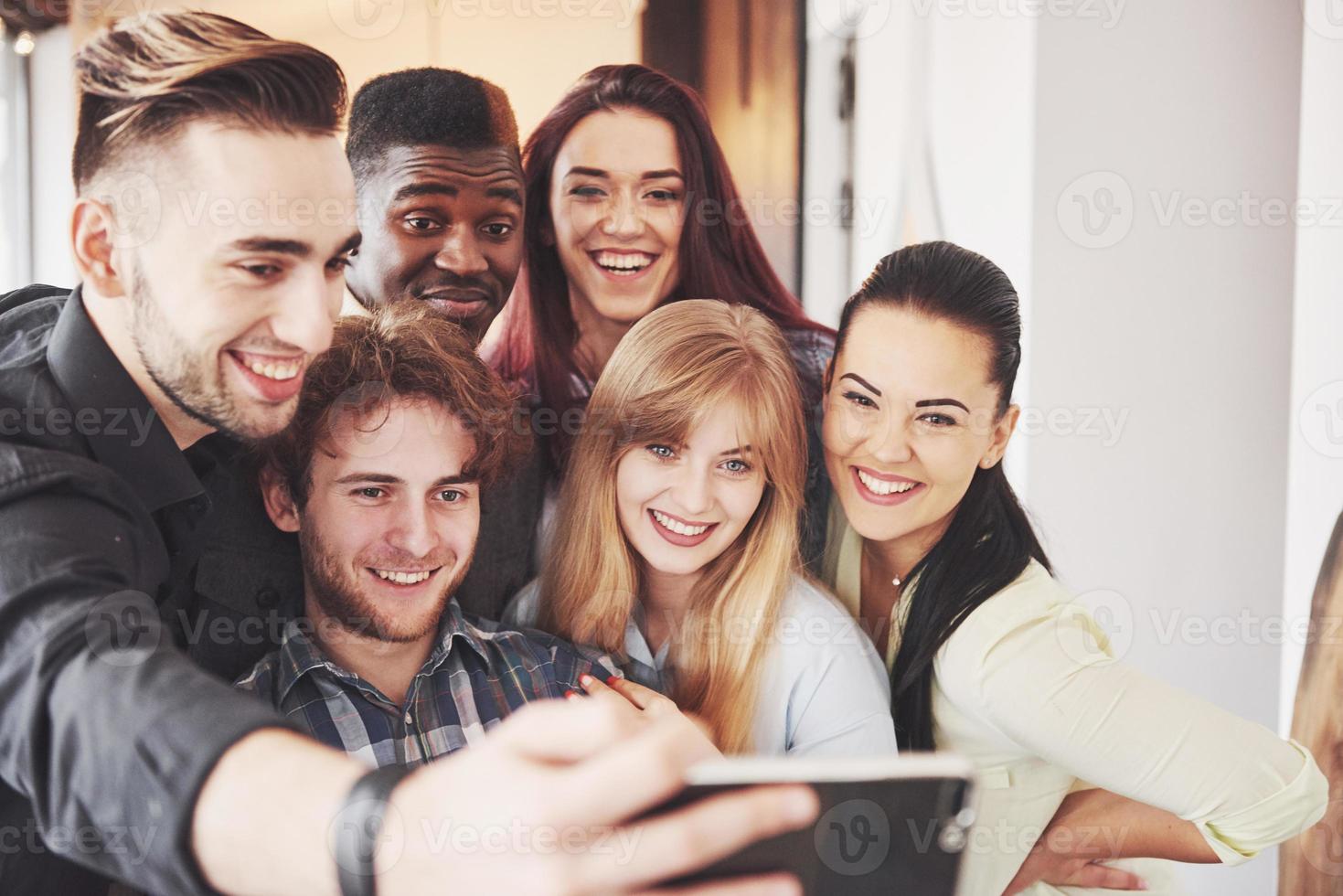 amici che si divertono al ristorante.due ragazzi e quattro ragazze che bevono facendo selfie, facendo segno di pace e ridendo. in primo piano donna in possesso di smart phone. tutti indossano abiti casual foto