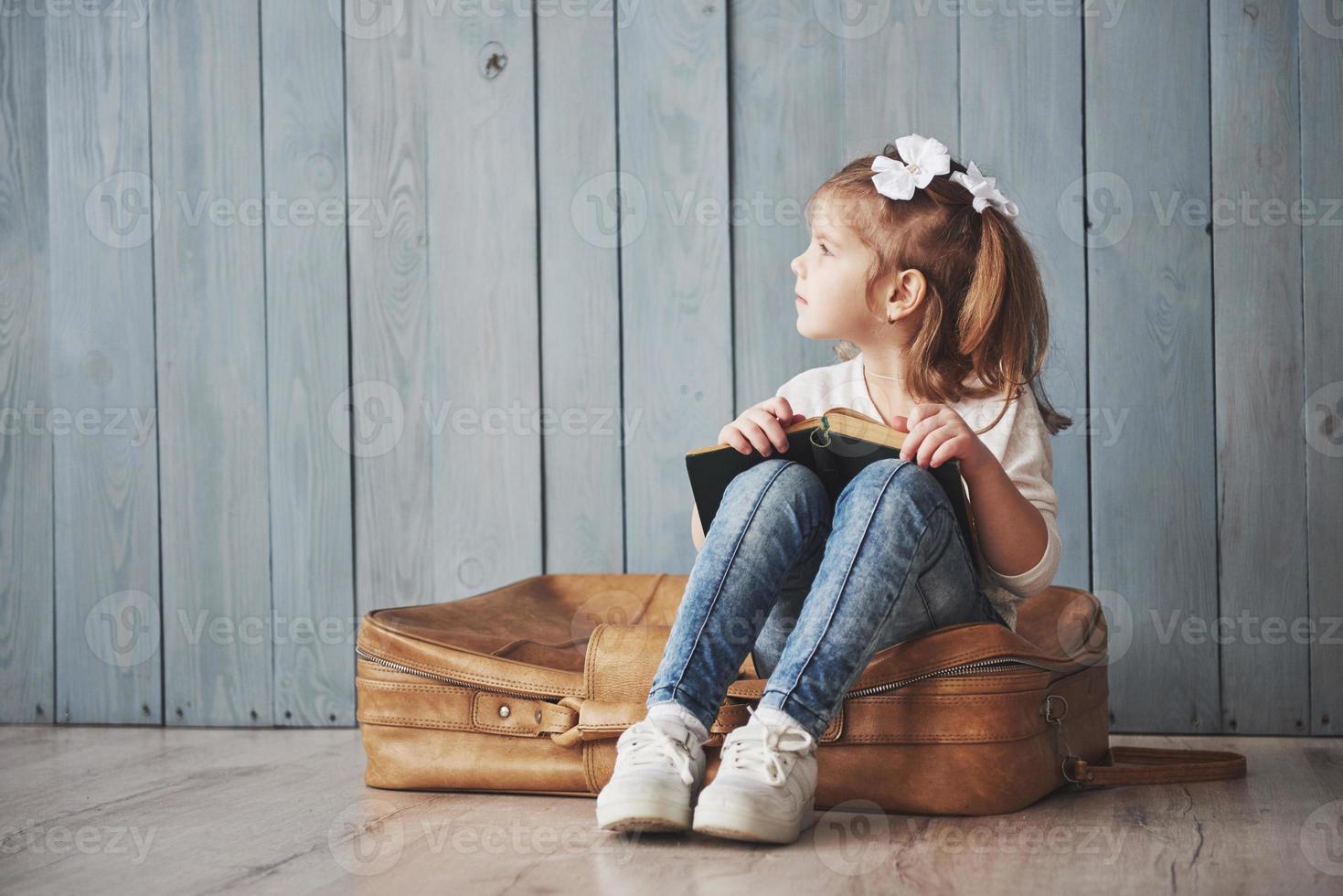 pronto per il grande viaggio. bambina felice che legge un libro interessante che trasporta una grande valigetta e sorride. concetto di viaggio, libertà e immaginazione foto