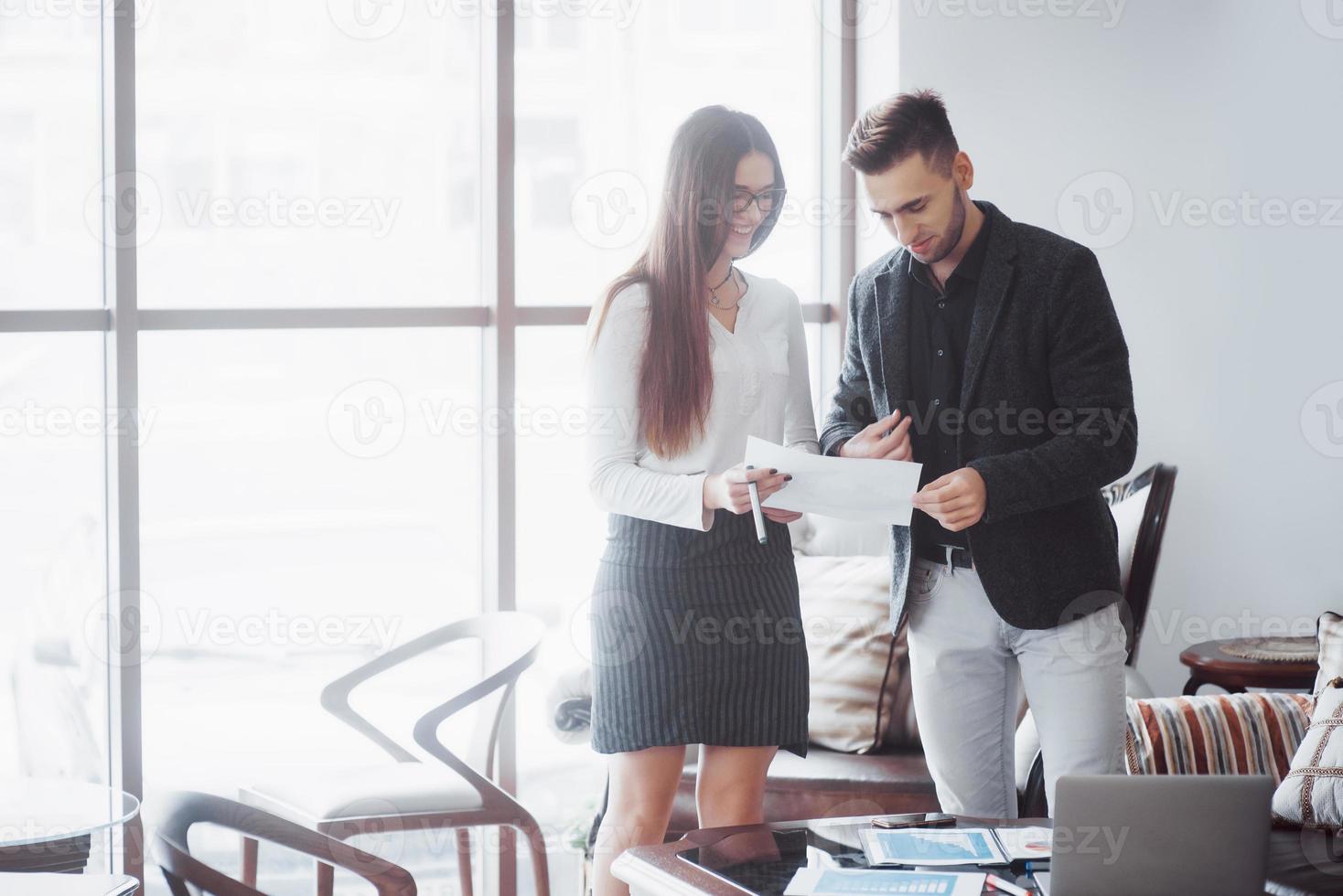 uomo d'affari e donna d'affari che studiano un grafico sul piatto e documenti cartacei alla finestra sullo sfondo dell'ufficio della città su un piano alto foto