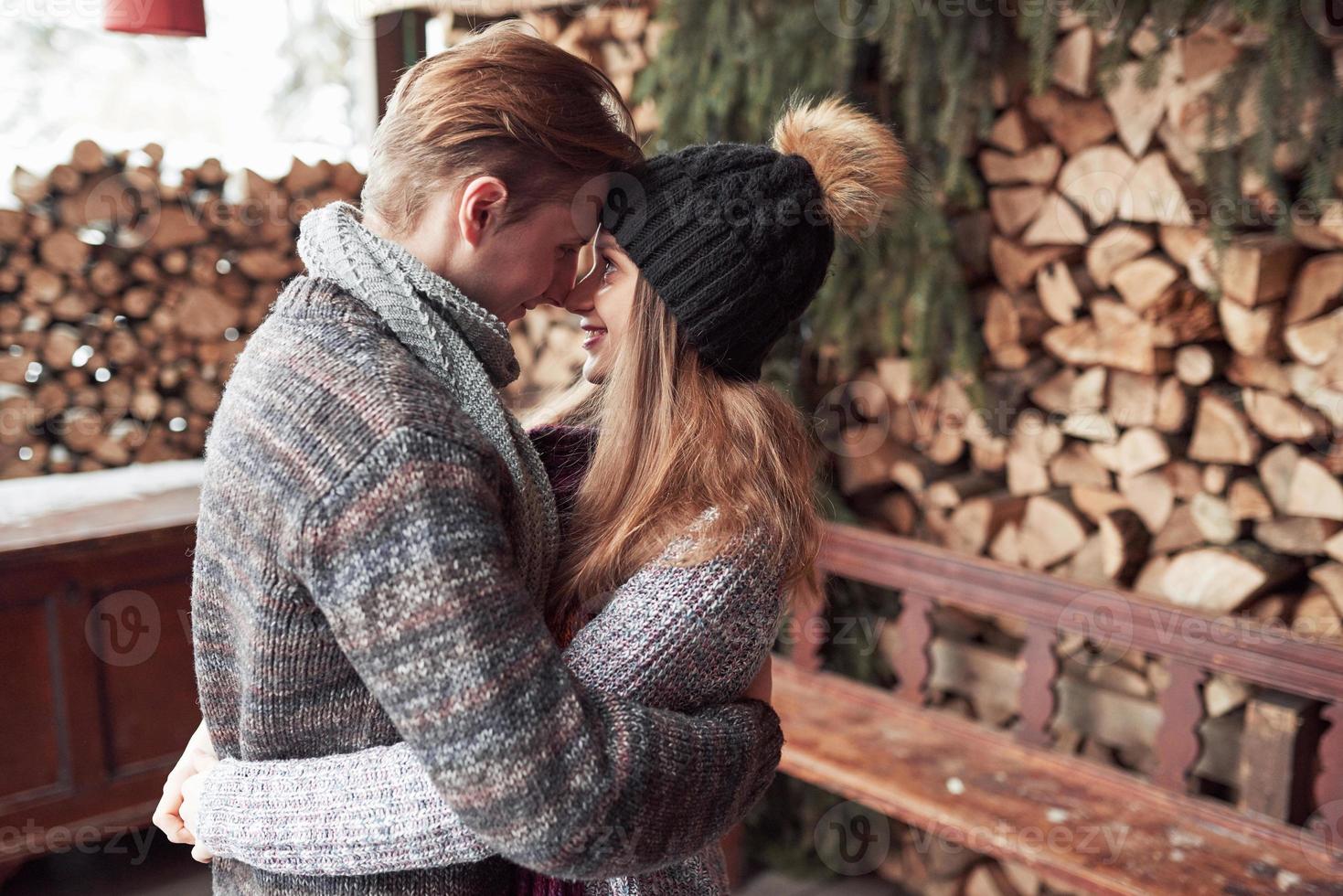 inverno, vacanza, coppia, natale e concetto di persone - uomo e donna sorridenti in cappelli e sciarpa che si abbracciano su una casa di campagna in legno e sullo sfondo della neve foto
