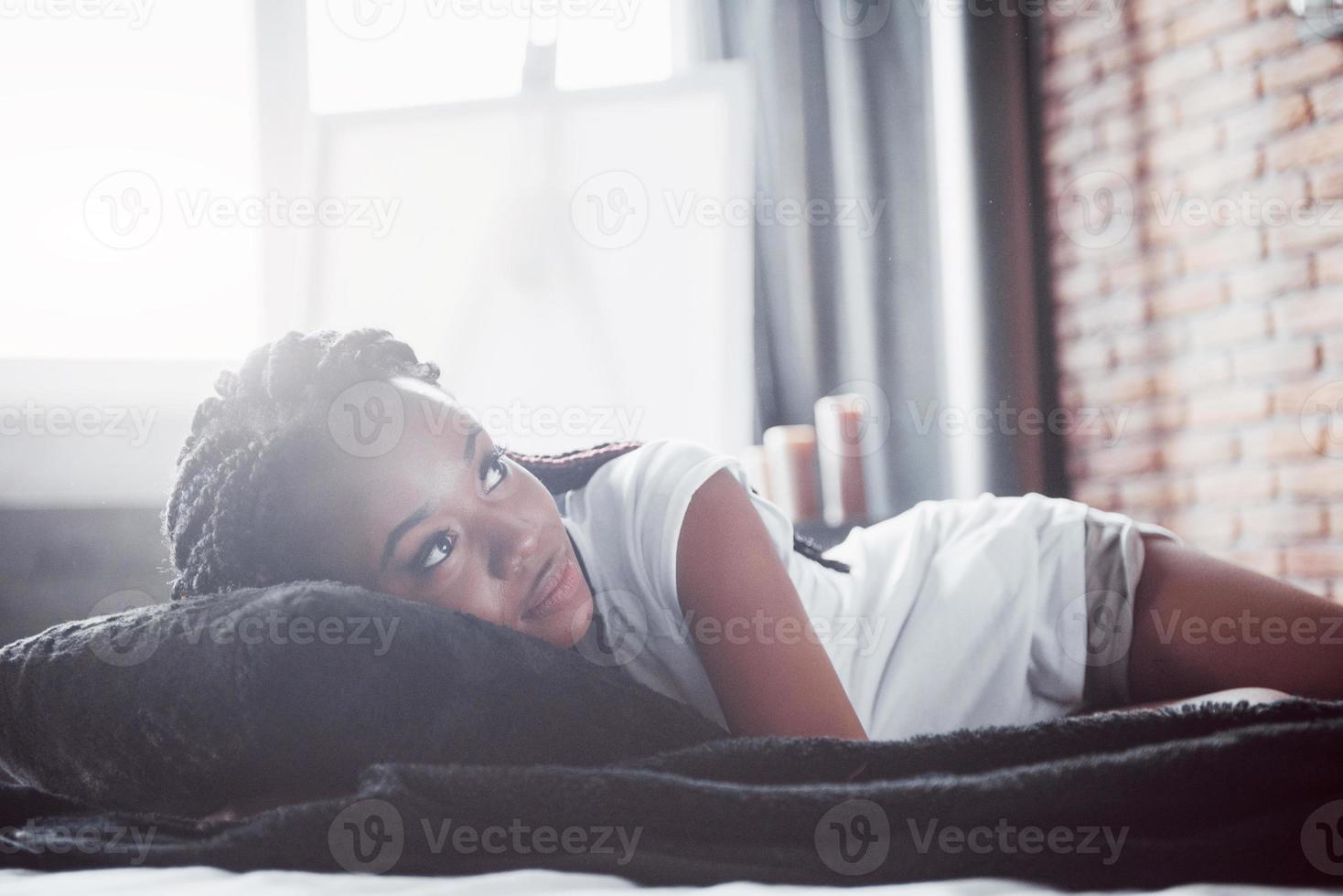 una bella donna africana giace e dorme a letto, in una camera da letto bianca. buongiorno, vacanza a casa. la ragazza indossa una t-shirt, codini in testa foto